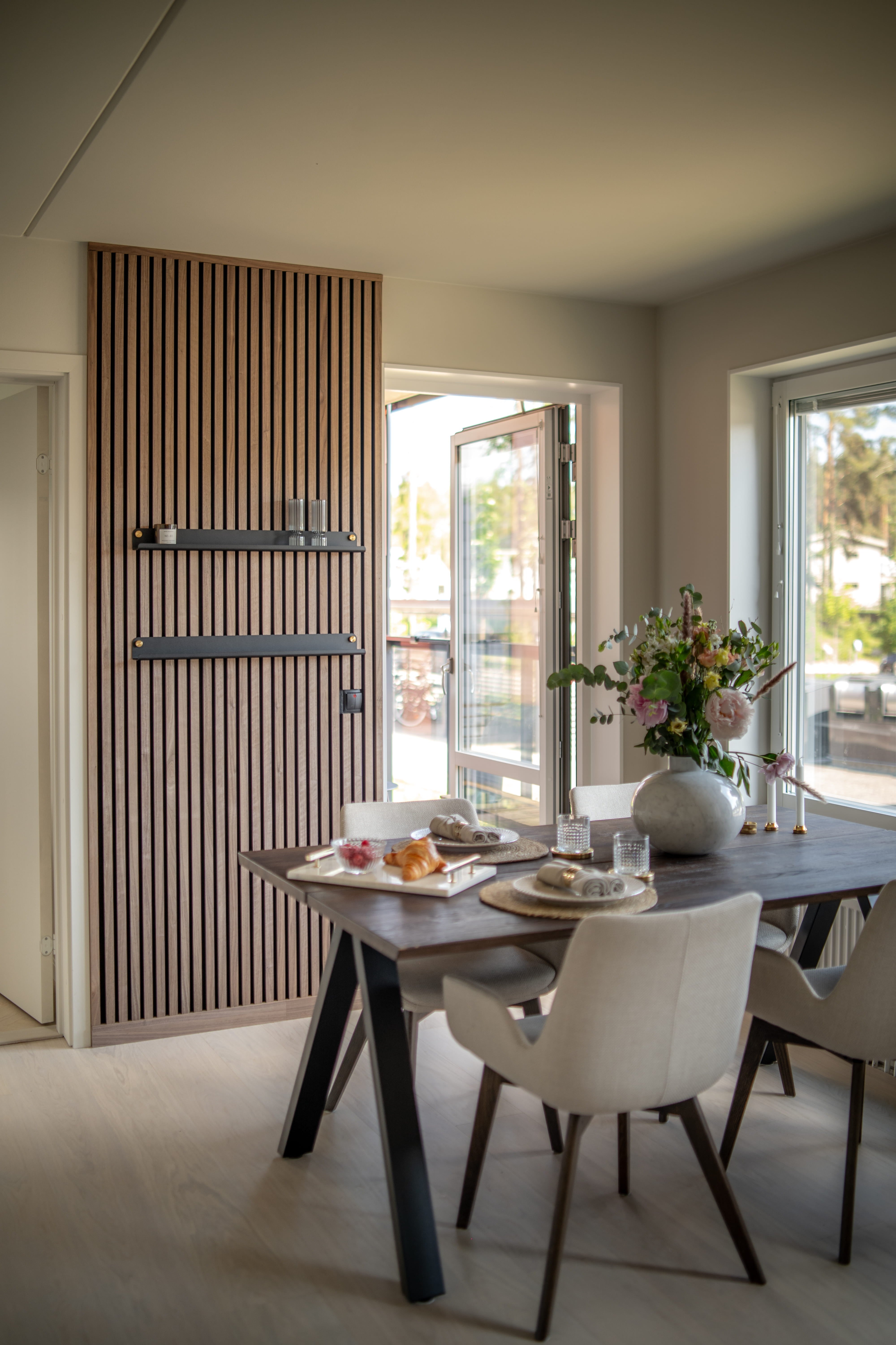Modern dining room with textured Akupanel acoustic wood slat wall, dark breakfast table, minimalist beige chairs, and a vase of flowers by large open windows.