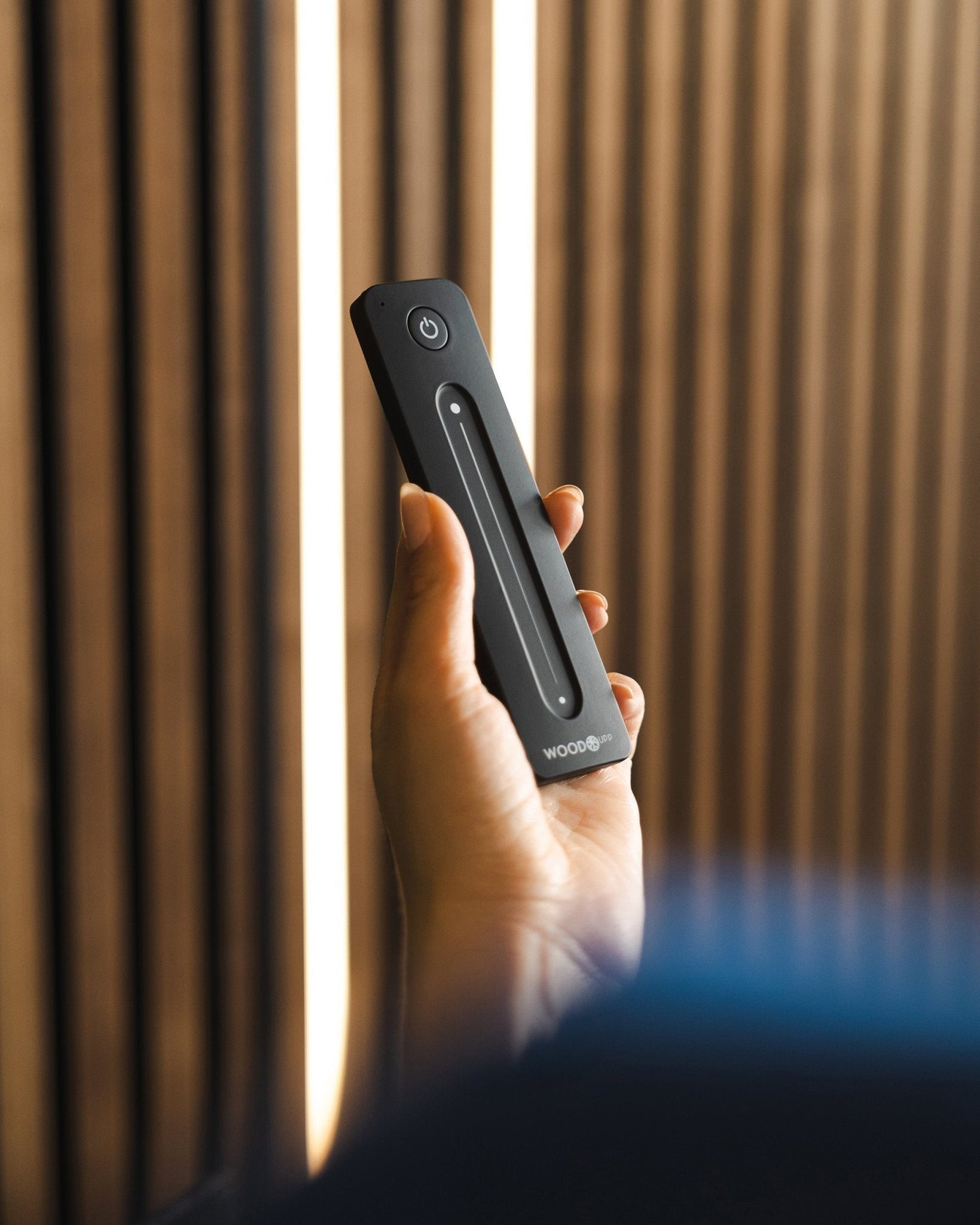 En hånd holder en black WoodUpp Lumi fjernkontroll til LED-lysstripe med av/på-knapp, plassert foran WoodUpp akustiske spilepaneler; kompatibel med Philips Hue.