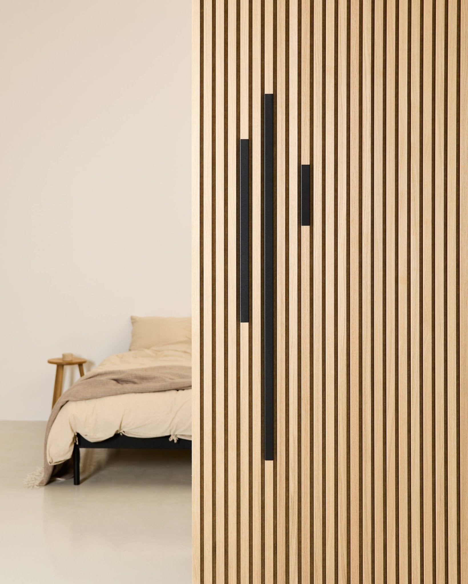 Minimalistisk soverom med en beige seng og et pledd, delvis skjermet av en 60 cm WoodUpp Akupanel skillevegg i Black aluminium fra Liva Cover-serien.