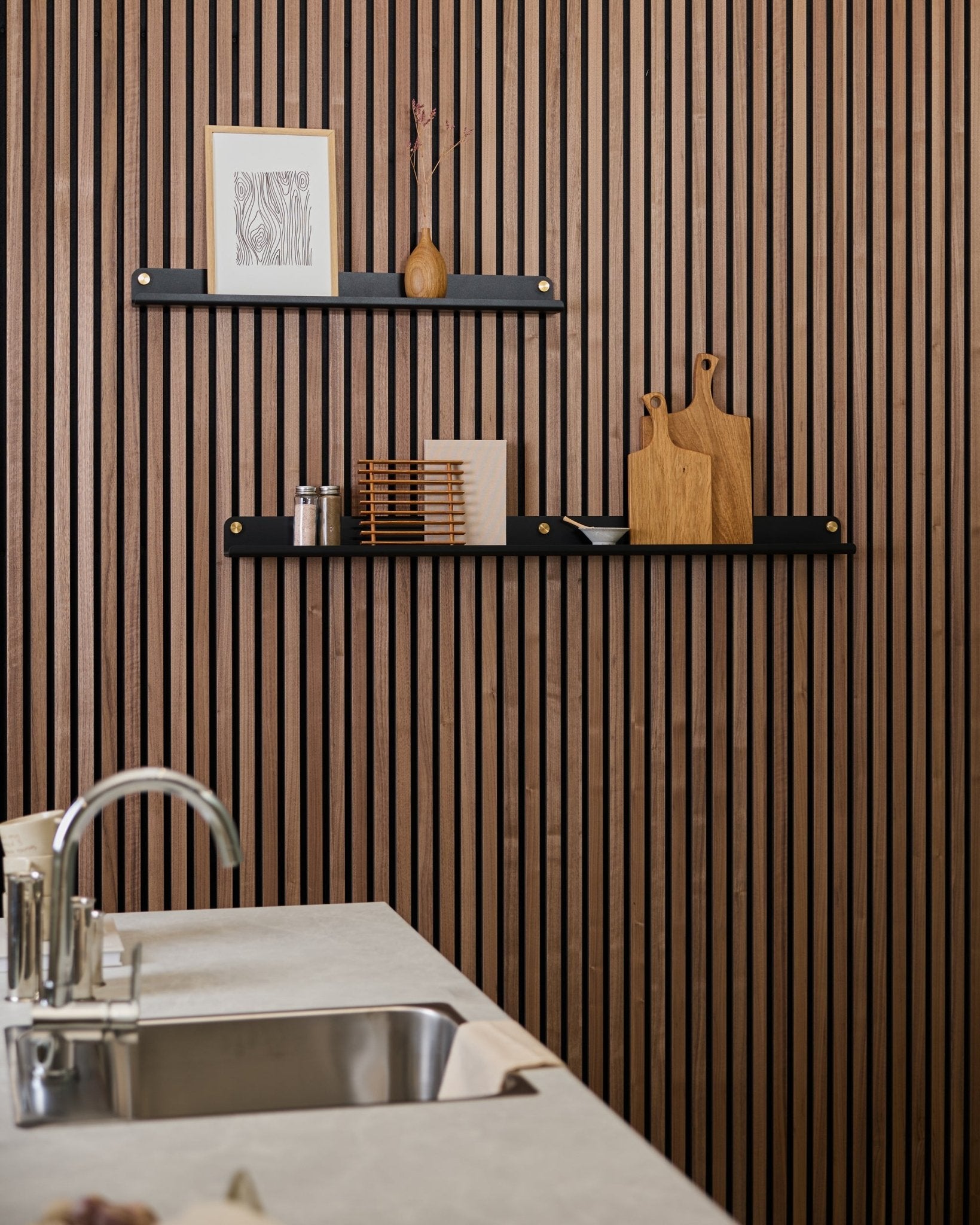 Minimalistisk kjøkken med en vertical Akupanel vegg med akustiske trespiler, black hyller, skjærefjøler, small dekorobjekter og en moderne kjøkkenbenk med vask foran.