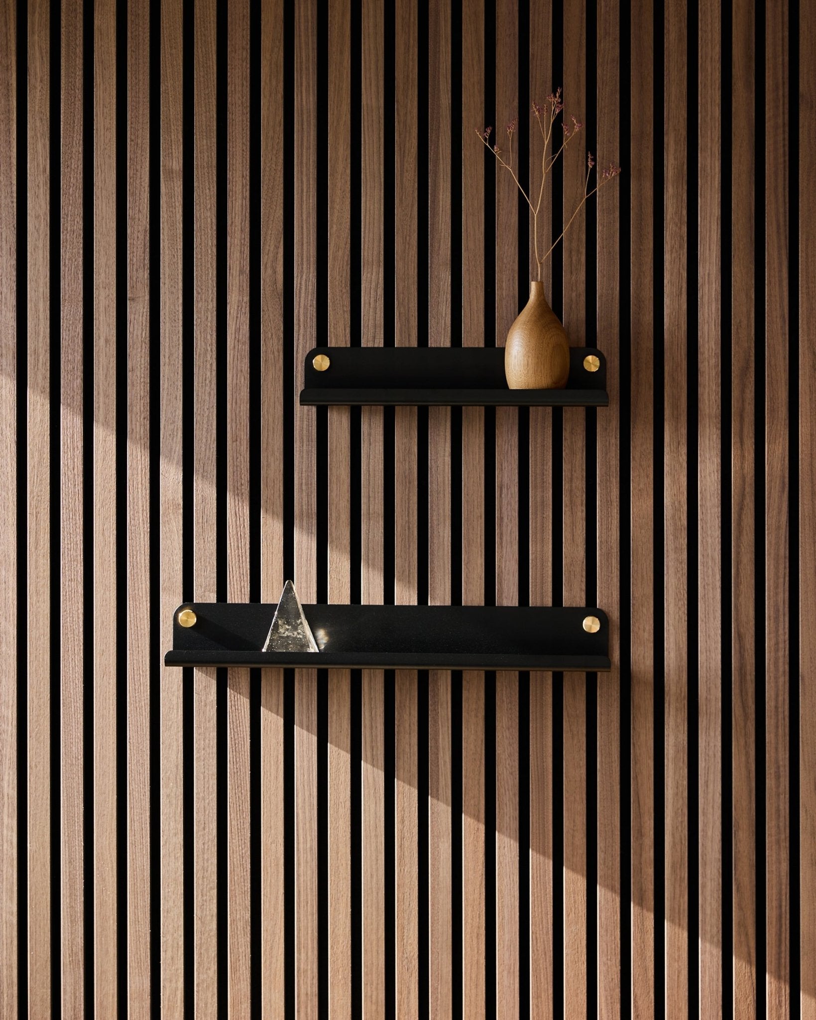 To black WoodUpp Norr smale hyller (35,9 cm) montert på et nordisk akustikkpanel med spiler; øverste hylle med en vase med tørkede grener, nederste med en pyramide i glass.