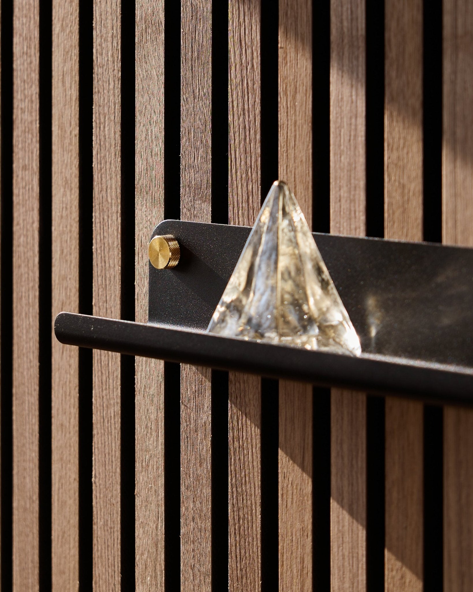 En glasspyramide står på WoodUpp Norr Narrow Shelf i black (51,9 cm), montert på en vertical Akustikkpanel og fremhevet av sollys.