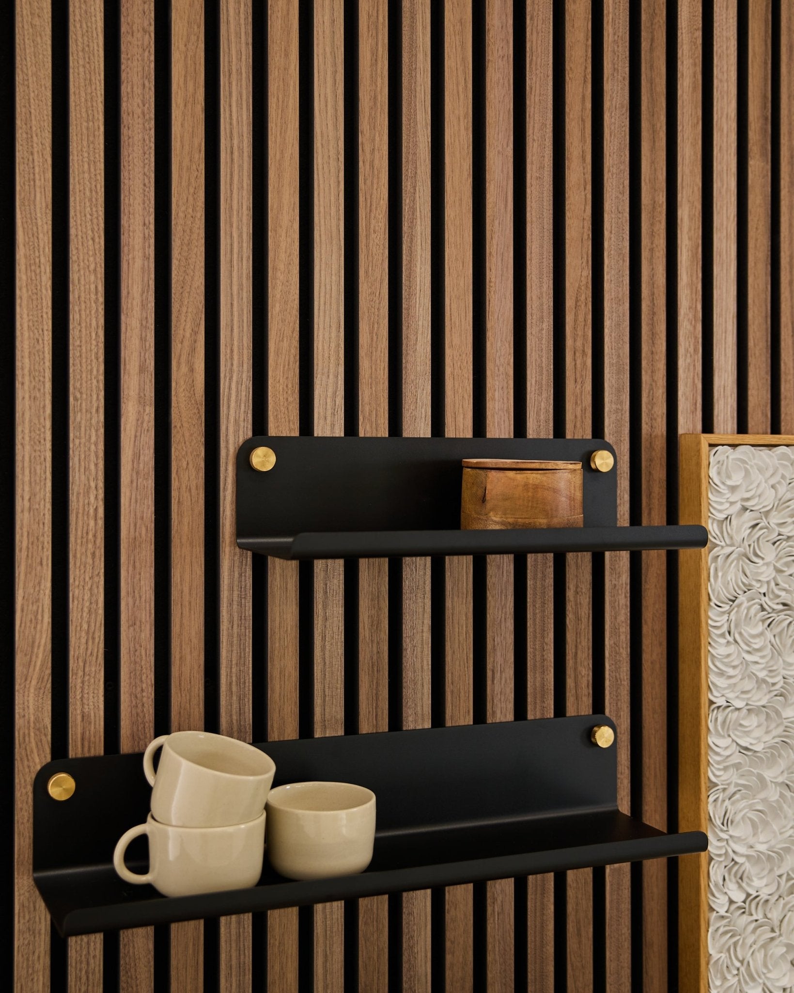To black Norr | Wide Shelves fra WoodUpp (35,9 cm) med gullfargede braketter montert på et Akupanel akustisk trepanel; den nederste hyllen har 3 beige krus, den øverste har en small treboks.