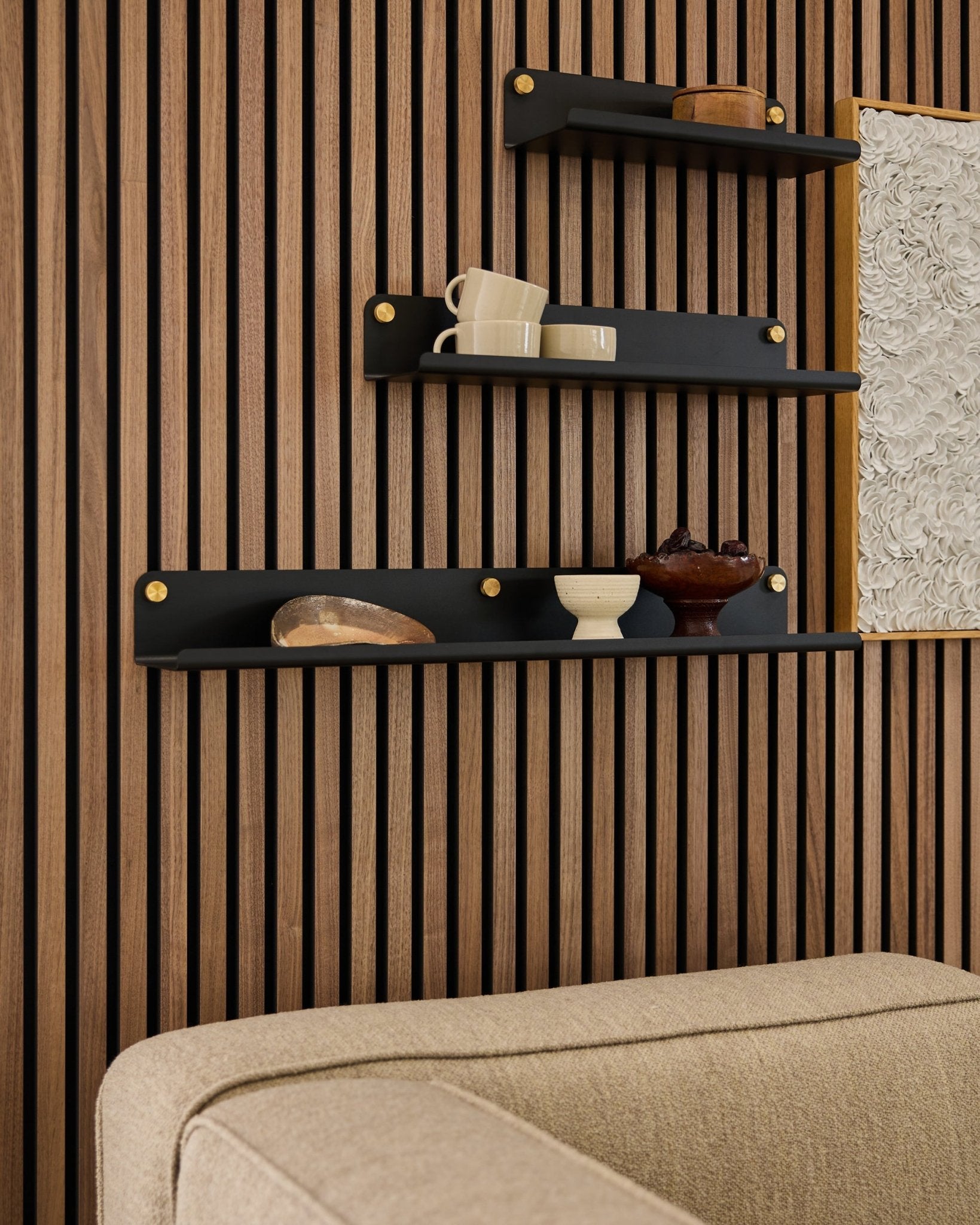 Tre black hyller, inkludert WoodUpp Norr Wide Shelf (75,9 cm), viser dekor på en Akustikkpanel-vegg med trespiler over en beige sofa; deler av et kunstverk er synlig.