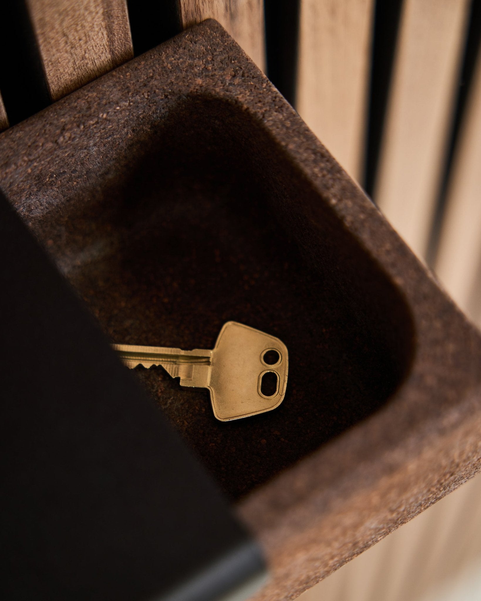 En brass nøkkel ligger i en Norr Key Tray/røkt kork, 35,9 cm fra WoodUpp, plassert på en brown beholder foran en bakgrunn av Akupanel akustiske trepaneler.