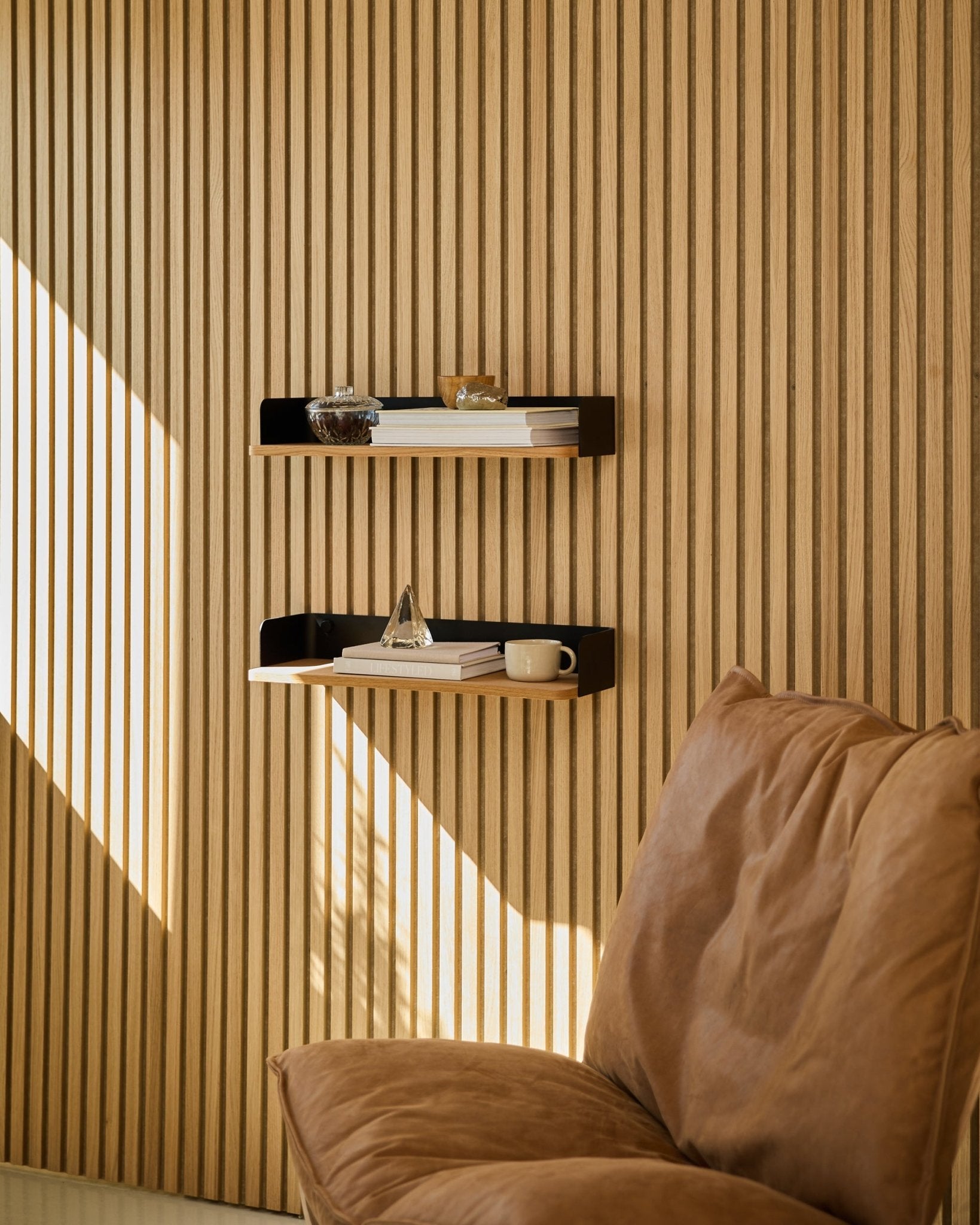 Brown stol ved siden av WoodUpp Linn Bookshelf i Natural Oak, med bøker og dekor arrangert i naturlig lys foran en nordisk Akustikkpanel spilevegg i tre.
