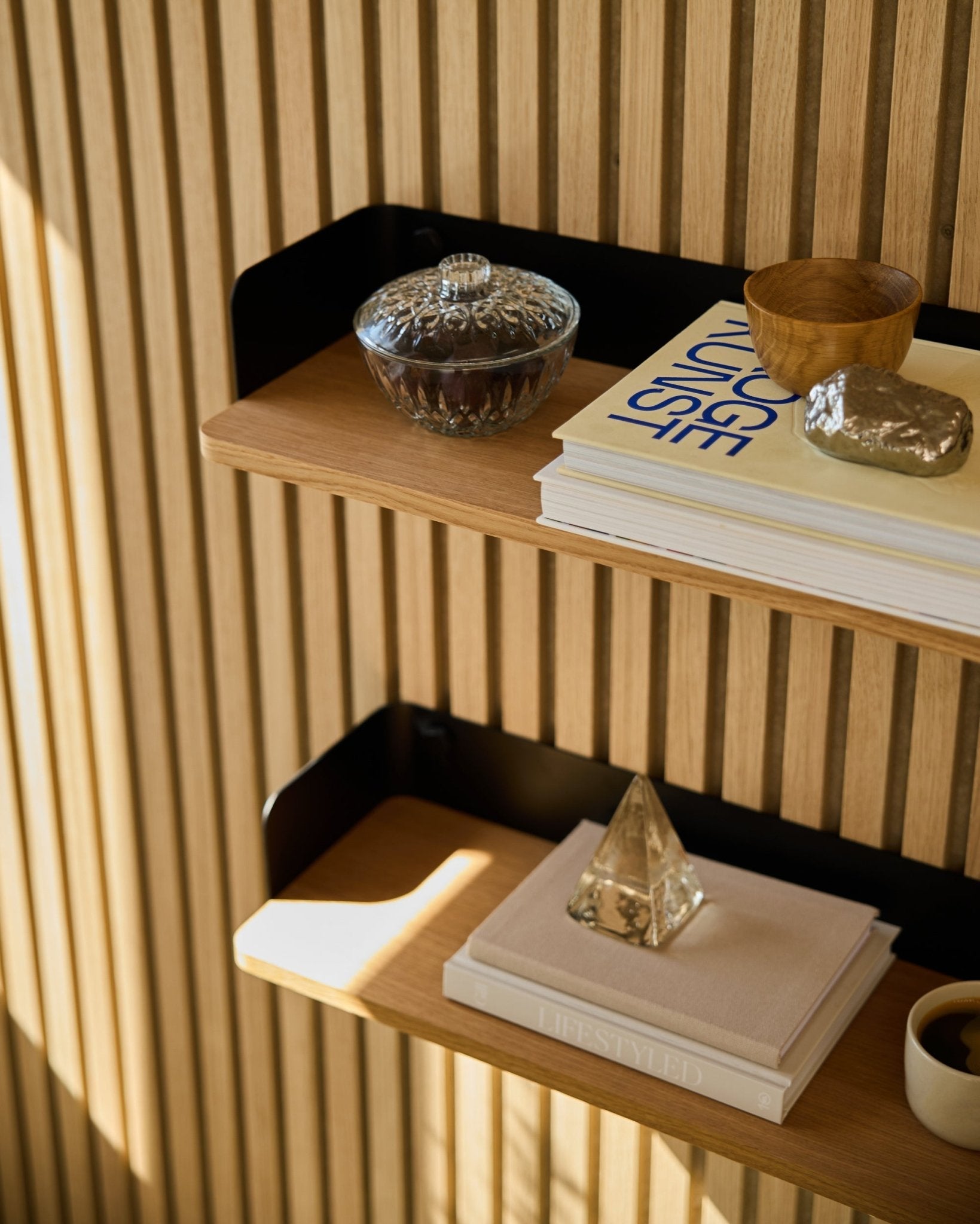 WoodUpp Linn Bookshelf i Natural Oak (55,9 cm) rommer en glasskrukke, kopp, gullfarget pyramide og bøker foran Akupanel akustiske trepaneler i sollys.
