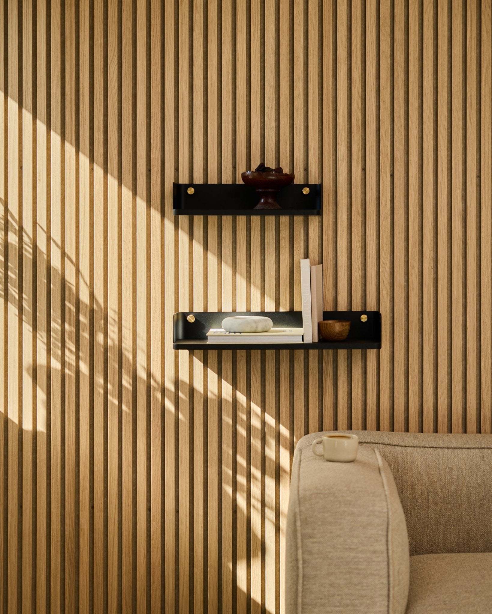 Sollys lyser opp en sofa i beige og WoodUpps bokhylle Linn i black oak, 55,9 cm, plassert mot en minimalistisk nordisk spilevegg Akustikkpanel.