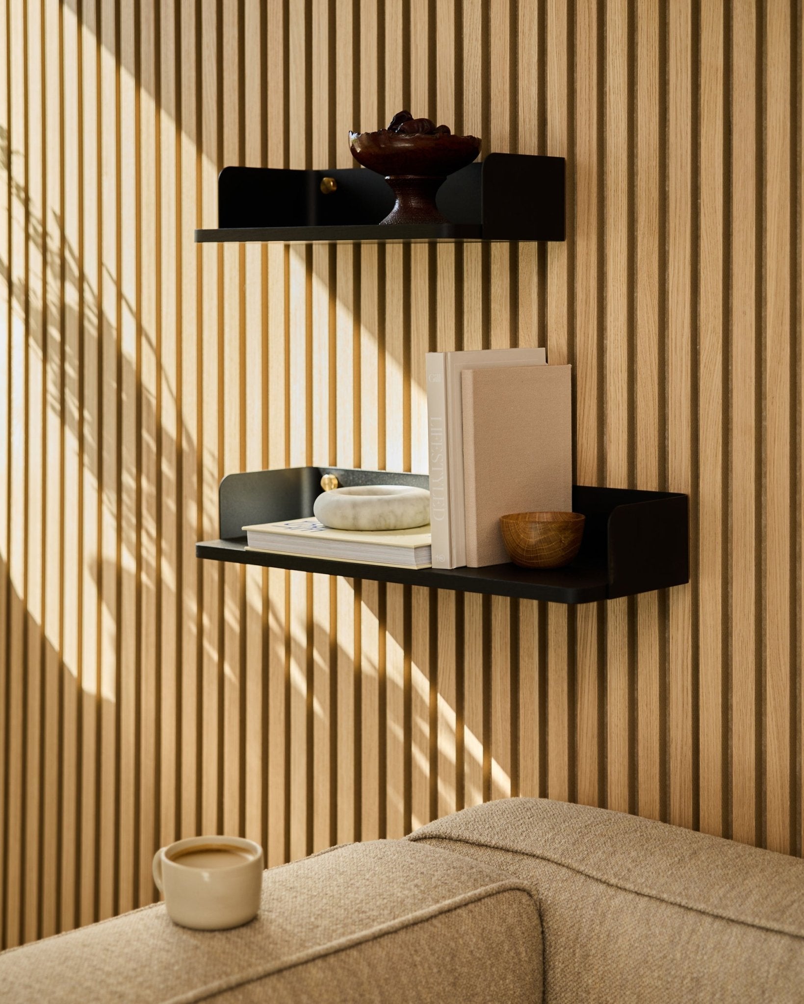 En black oak WoodUpp-bokhylle (55,9 cm) viser frem bøker, dekor og en skål på en vegg med akustiske trepaneler; en sofa i beige og et krus står i forgrunnen.