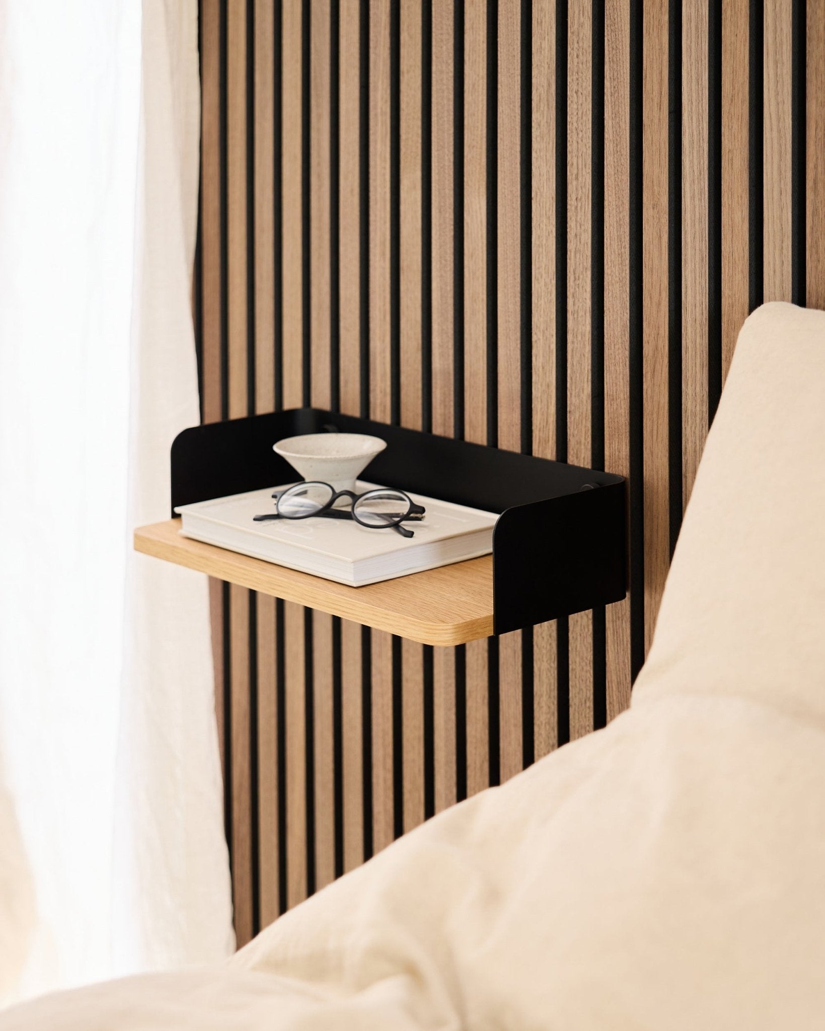 Linn Bookshelf fra WoodUpp i natural oak (39,9 cm) med en bok, briller og et glass ved siden av en seng mot en vegg med Akupanel akustiske trepaneler.