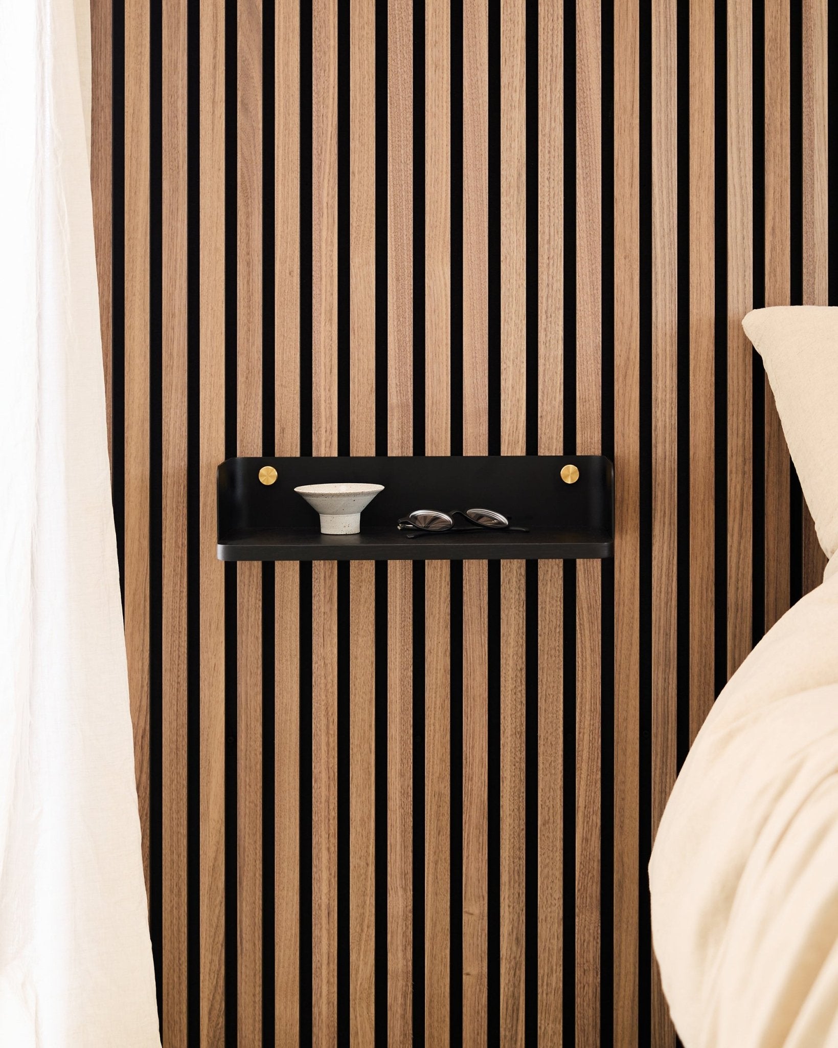 Linn Bokhyllen i Black Oak fra WoodUpp er montert på et nordisk akustisk spilepanel, og har en skål og glass ved siden av en seng med beige sengetøy.