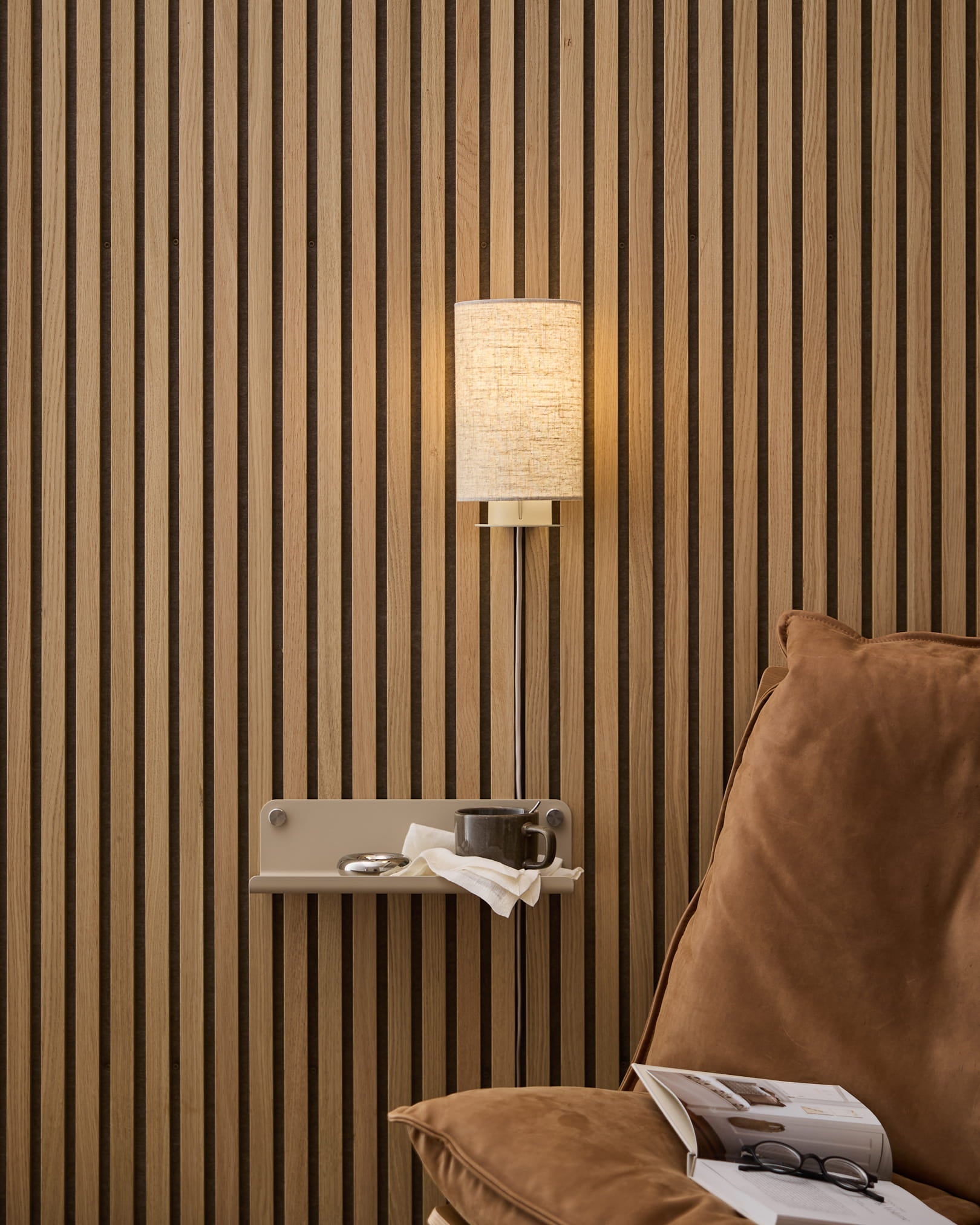 En beige lin-bomull WoodUpp Feya vegglampe montert over en lun lenestol og et sidebord med bok og kopp, satt mot en nordisk trespilet Akustikkpanel.