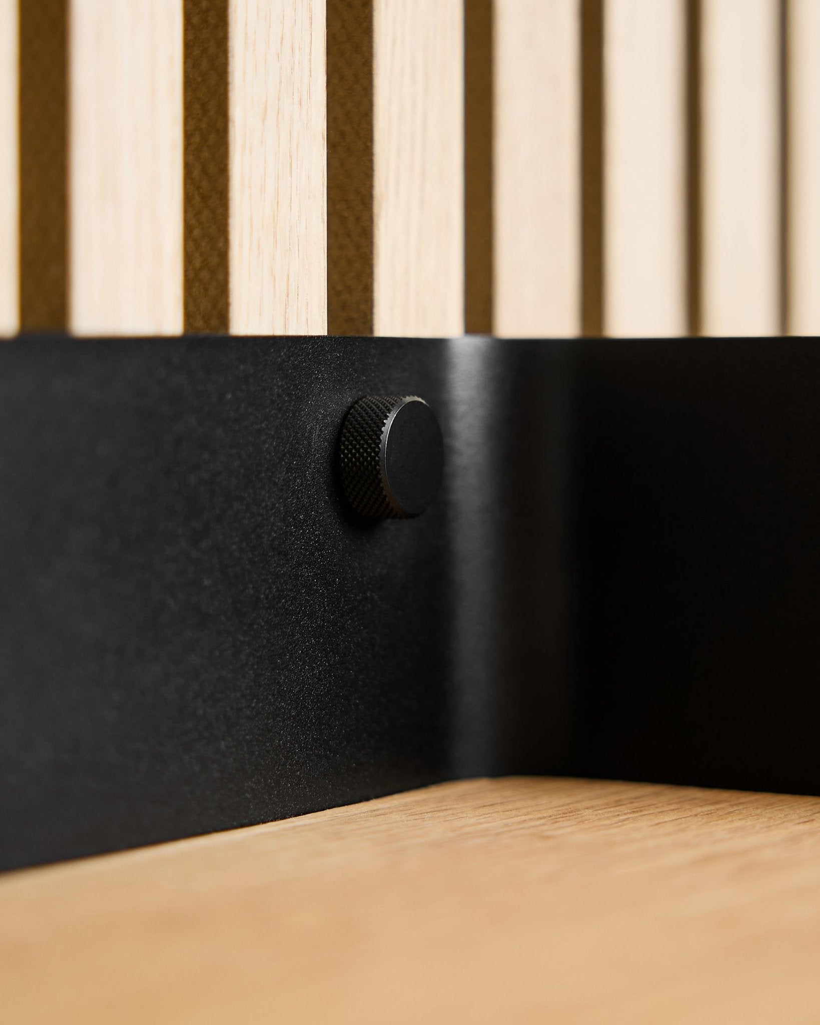 Nærbilde av to black WoodUpp Finger-skruer (M5x10 mm, 20x8,8) med brass skruer, plassert på en nordisk akustisk spilevegg i tre og en lys treoverflate.