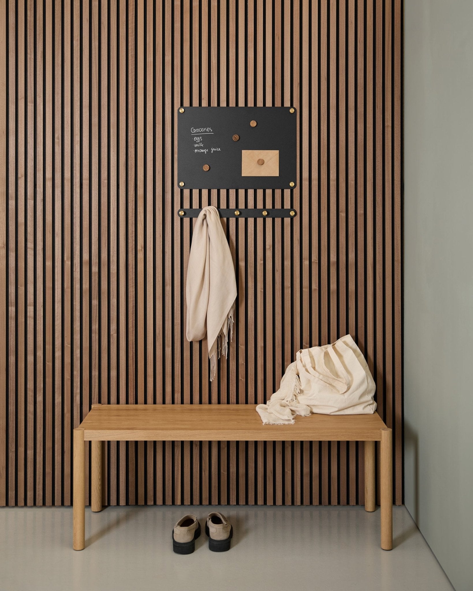 Minimalistisk entré med en trebenk, to skjerf, sandaler og en small WoodUpp Norr magnettavle på et Akupanel akustisk spilepanel i tre.