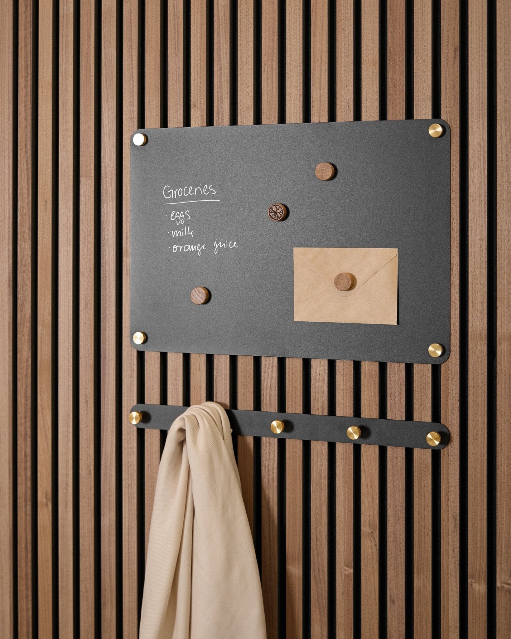 En small black Norr magnettavle fra WoodUpp holder en handleliste, en konvolutt, magneter og beige stoff på kroker, montert på en Akupanel Akustikkpanel.