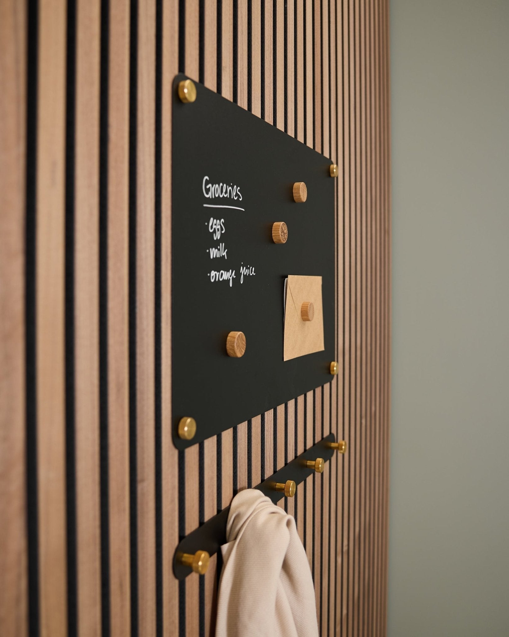 En small black Norr magnettavle fra WoodUpp montert på et Akupanel akustisk trepanel viser en handleliste og et notat, med kroker under som holder et lyst stoff.