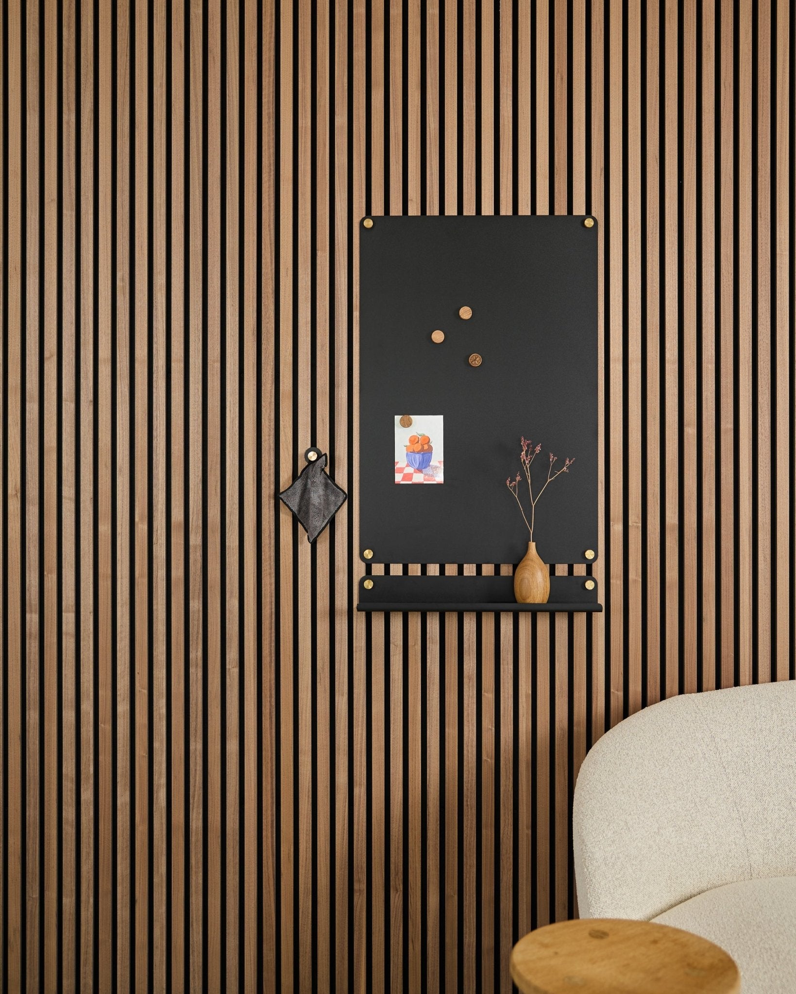 En large black Norr Magnettavle fra WoodUpp, laget for akustikkpaneler, henger ved siden av en hvit polstret stol med notater, en vase og en liten dekorasjon.