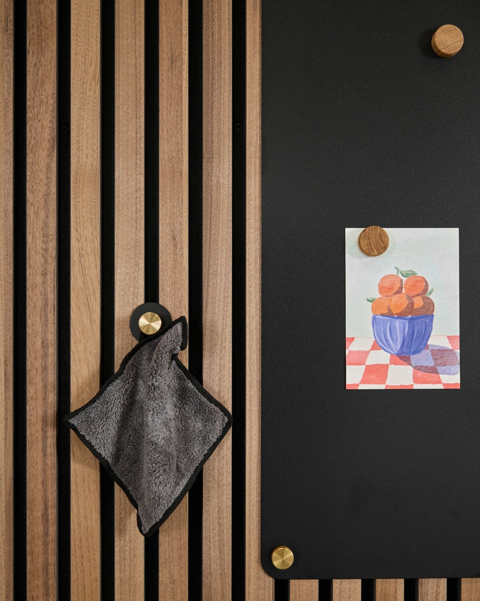 Et small håndkle henger på et nordisk akustisk spilepanel ved siden av en black tavle med en tegning av appelsiner i en bolle, festet med to gullfargede magneter.