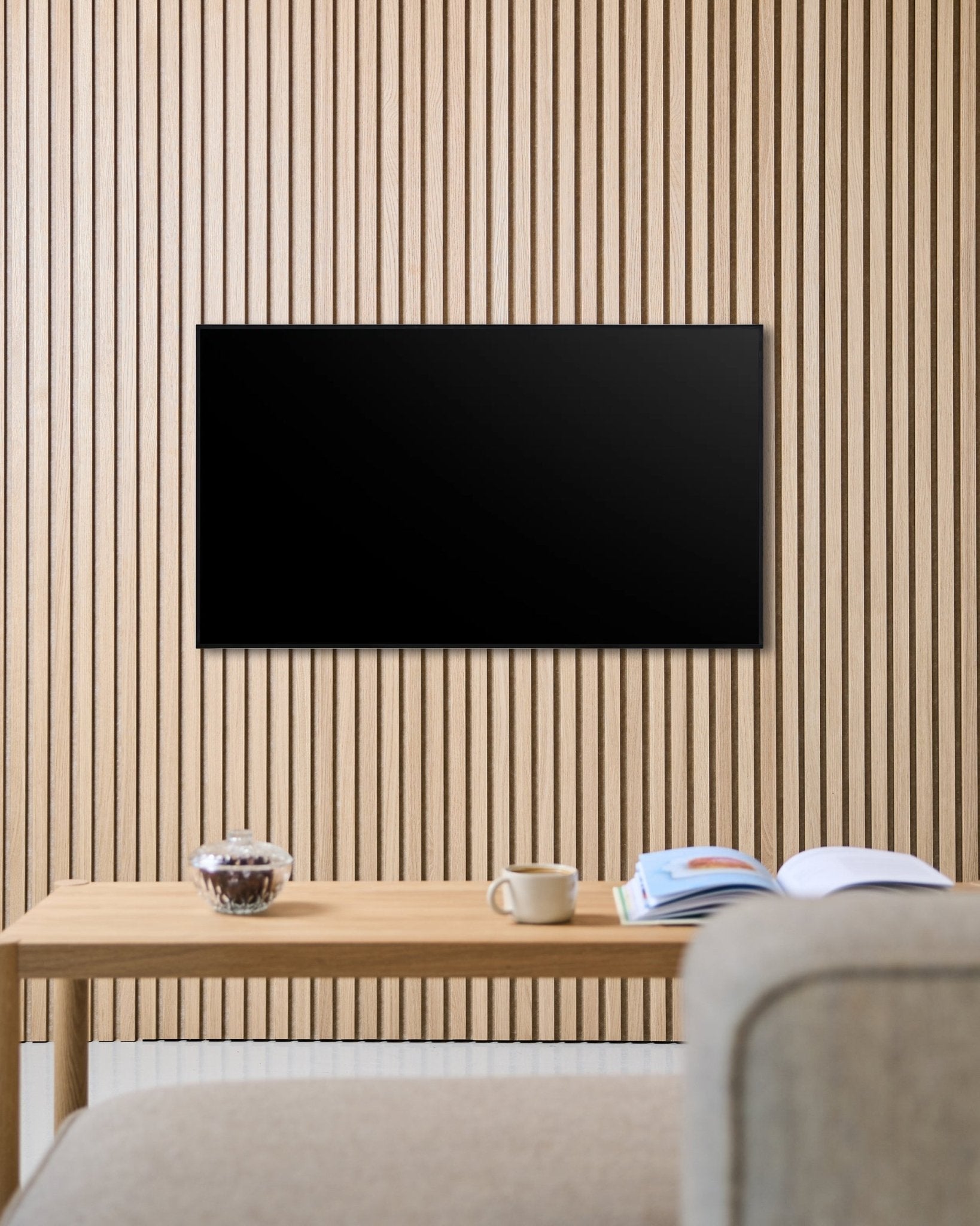 Et Fjell TV-feste (small) fra WoodUpp fester en flatskjerm til en nordisk Akupanel akustikkvegg med trespiler, over et bord med kopp, skål og et åpent magasin ved siden av en beige sofa.