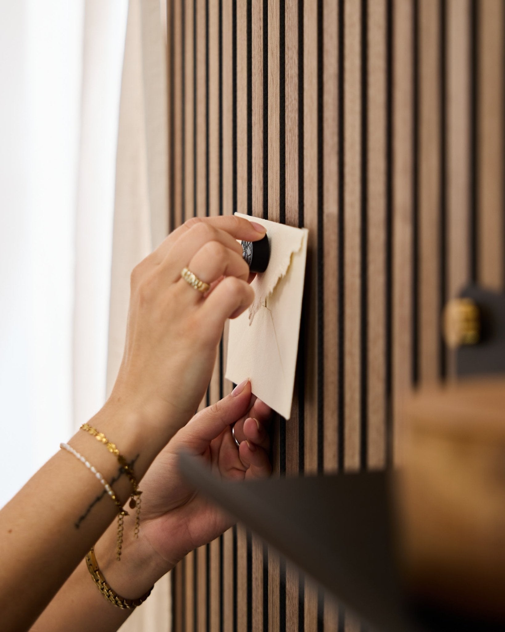 En person med gullarmbånd og ringer fester en konvolutt til et WoodUpp akustisk spilepanel i eik med Norr Poster Magnet-settet i Black Oak.