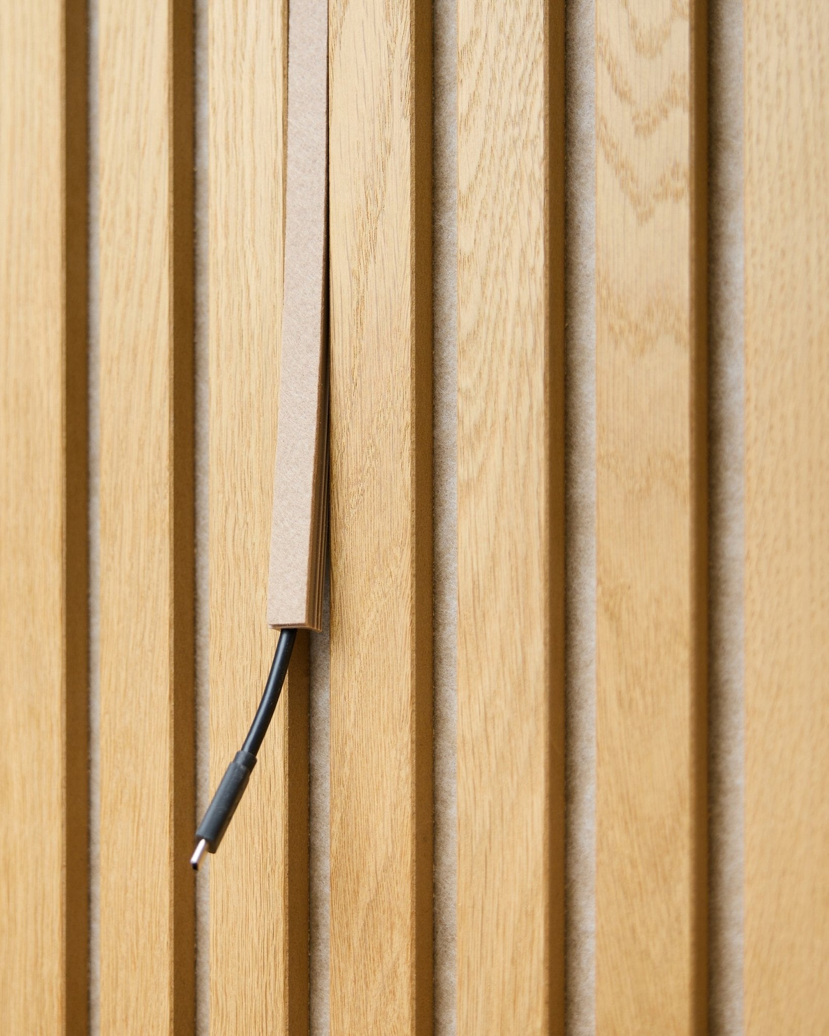 En black kabel med kabelhåndtering er delvis skjult i et WoodUpp Dupps Beige Pop-on Cable Cover, montert på vertical Akupanel akustiske trespiler.