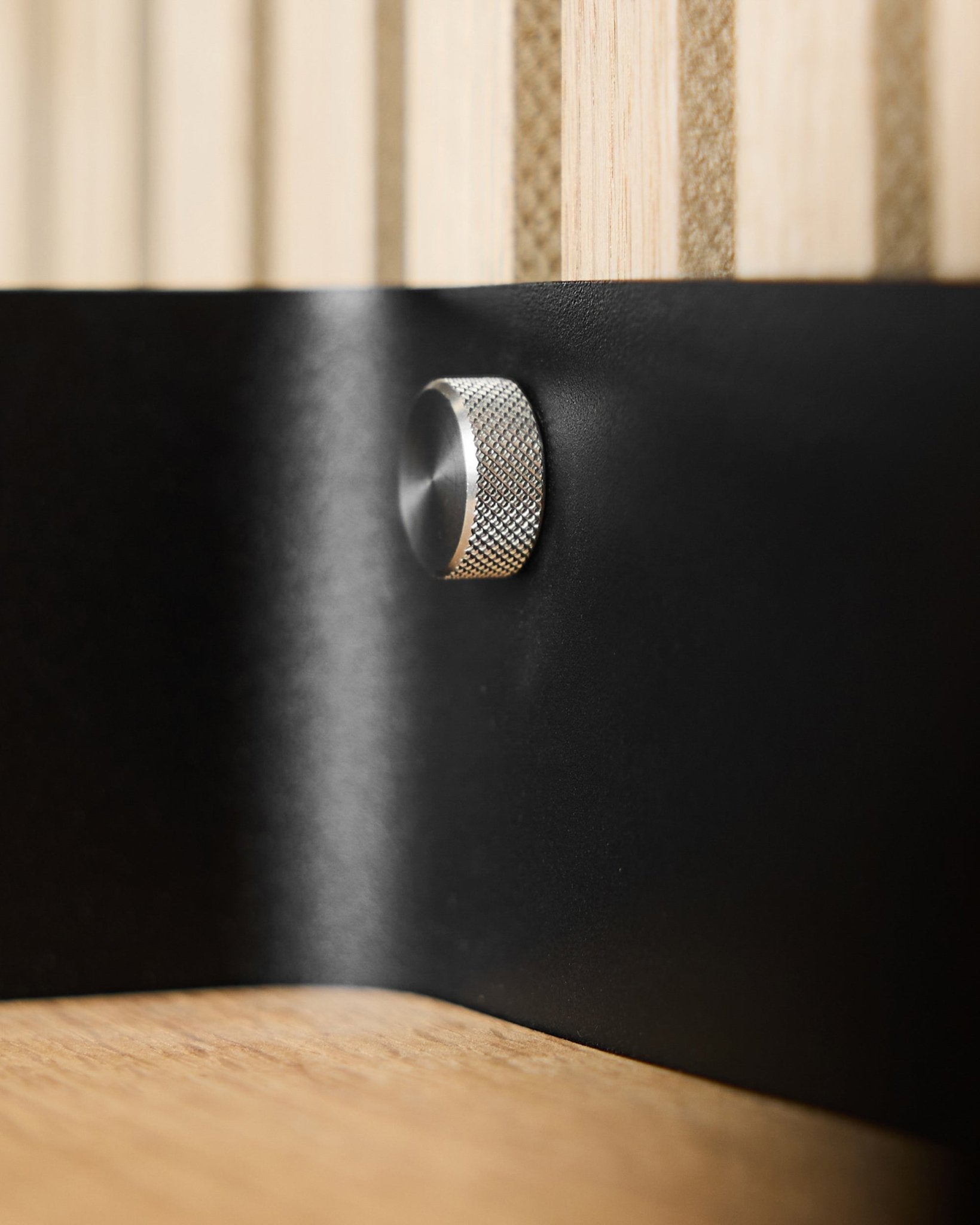 Nærbilde av to WoodUpp finger-skruer (M5x10 mm, 20x8.8 rustfritt steel) på en black metalloverflate med Akupanel akustiske trelameller i bakgrunnen.