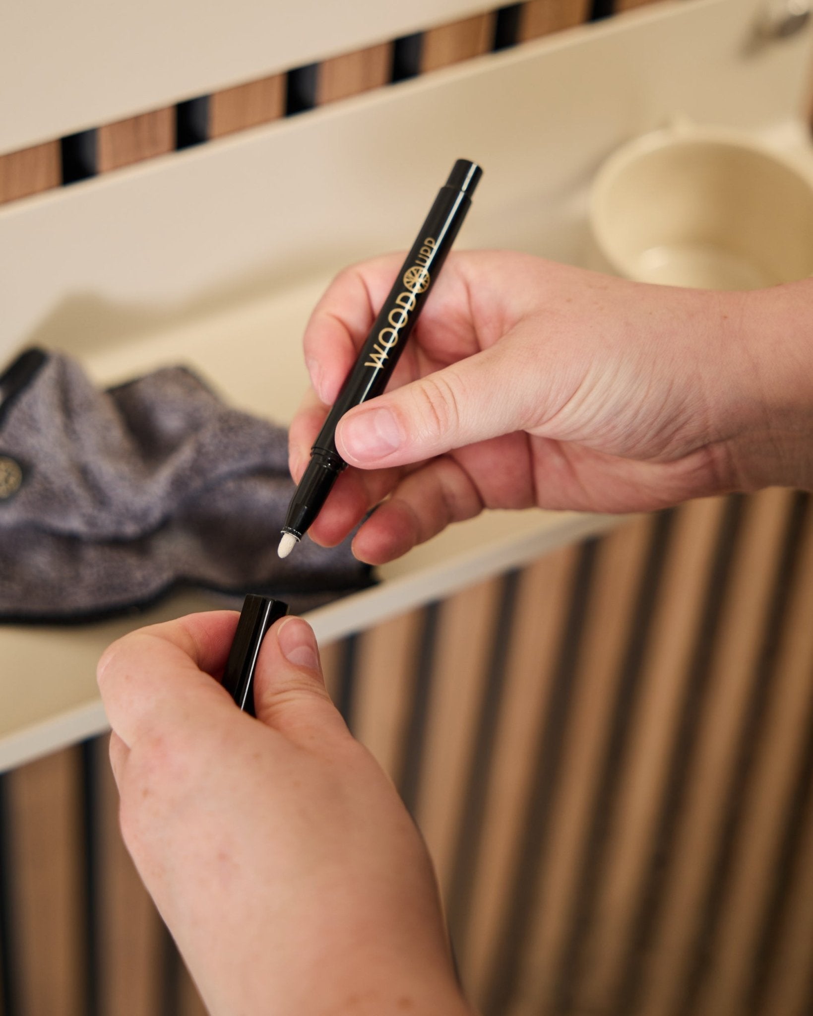 En person holder en åpen WoodUpp Norr | Blackboard set tusj ved siden av en mikrofiberklut og en beige kopp, produkter laget for bruk med akustiske trepaneler.