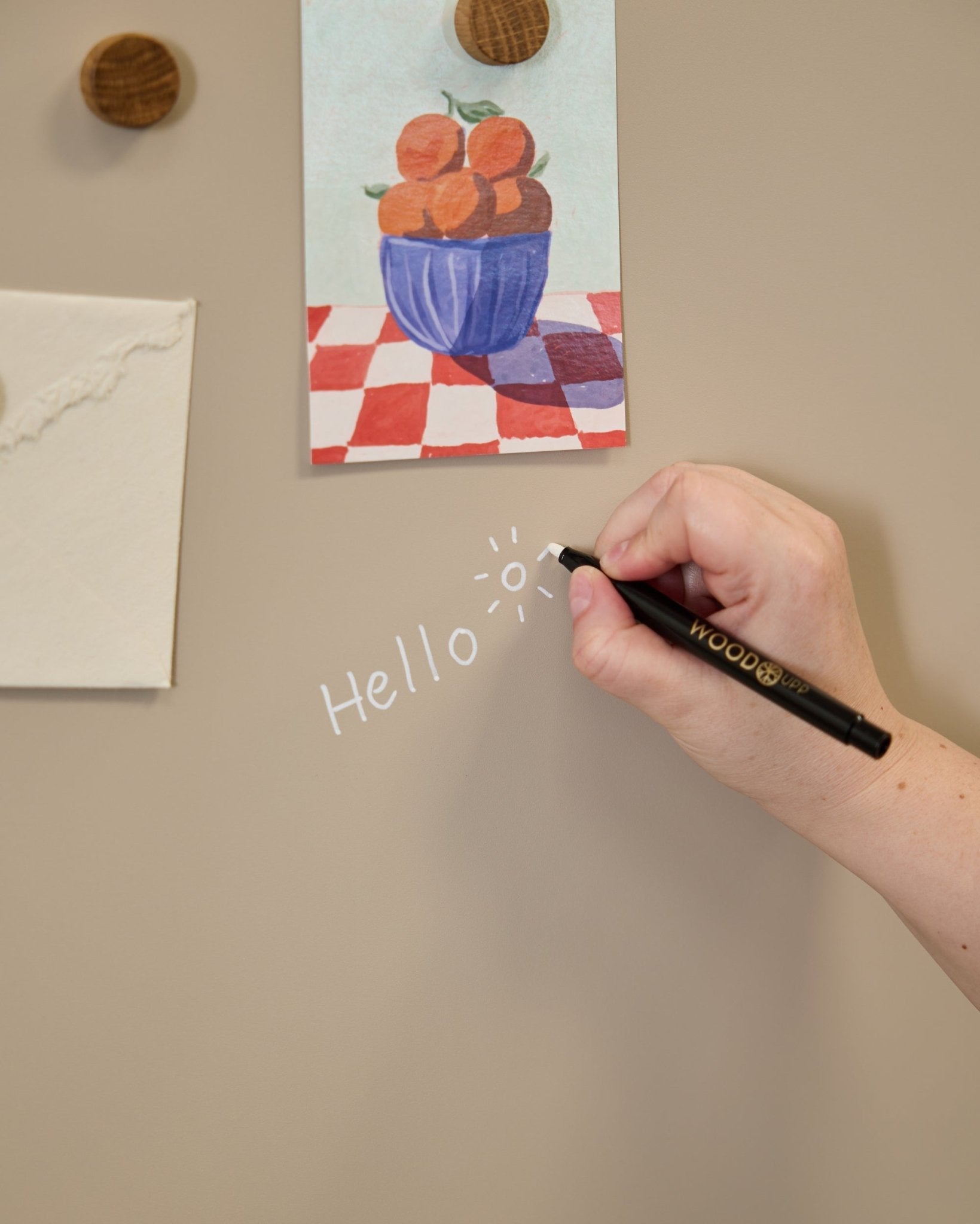 En hånd skriver hello med en soltegning på WoodUpp Norr Blackboard, plassert ved siden av kunst festet med runde magneter på en Akustikkpanel.