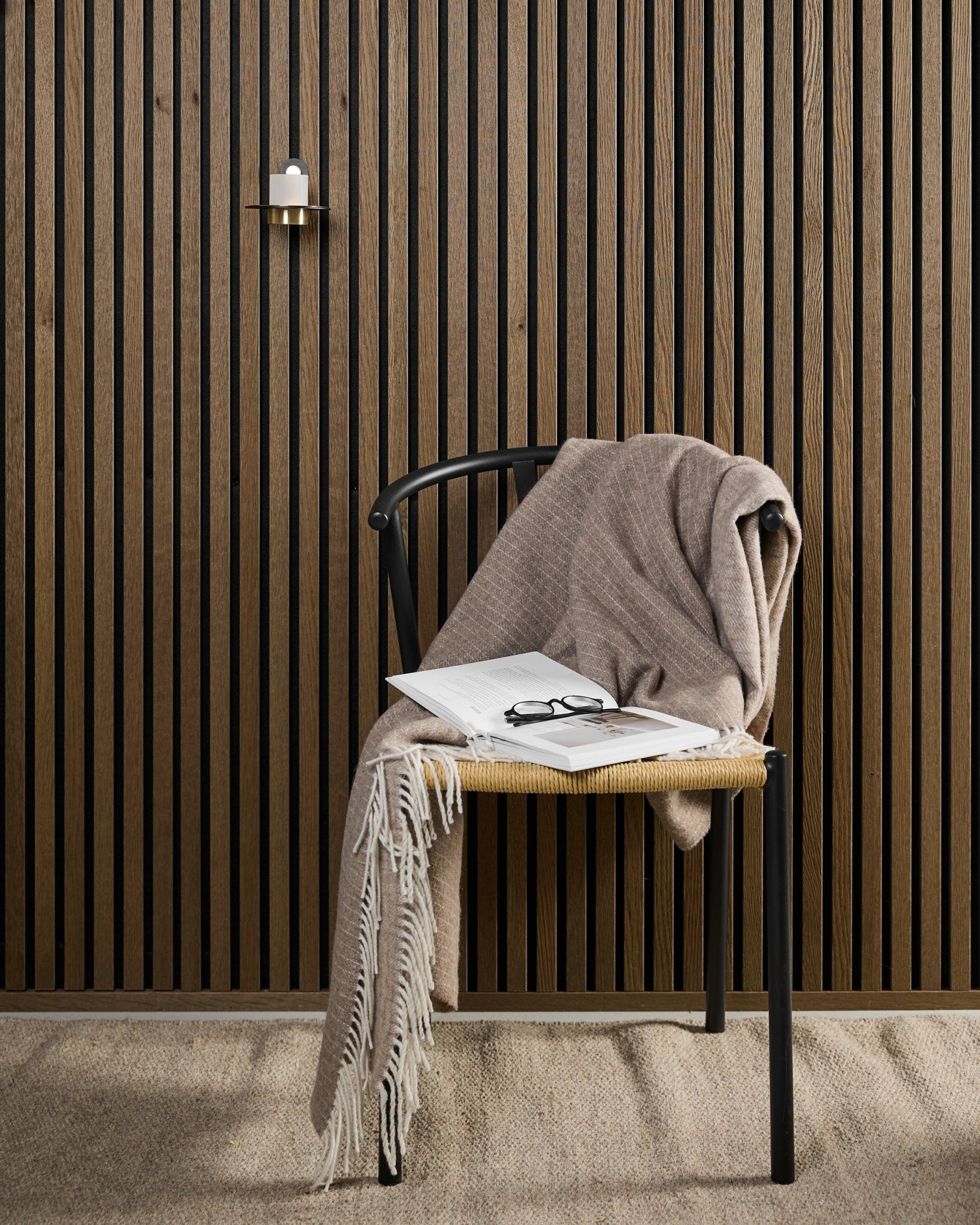 En moderne stol med et beige pledd, en åpen bok og briller står ved siden av et Akupanel akustisk trepanel og en black og brass WoodUpp Norr Candle Holder.