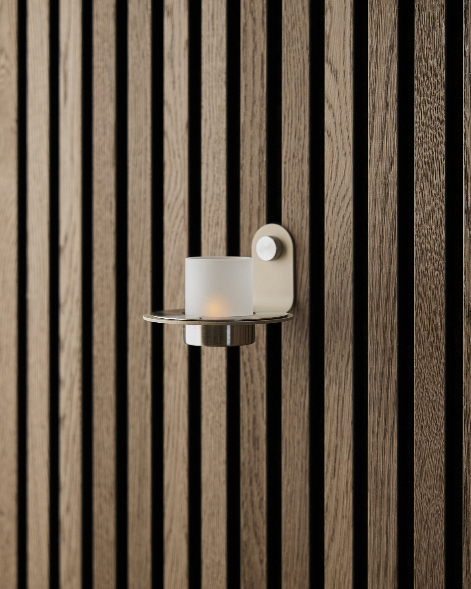 Norr Candle Holder i beige rustfritt steel fra WoodUpp, vist på en beige Akupanel Akustikkpanel-vegg.