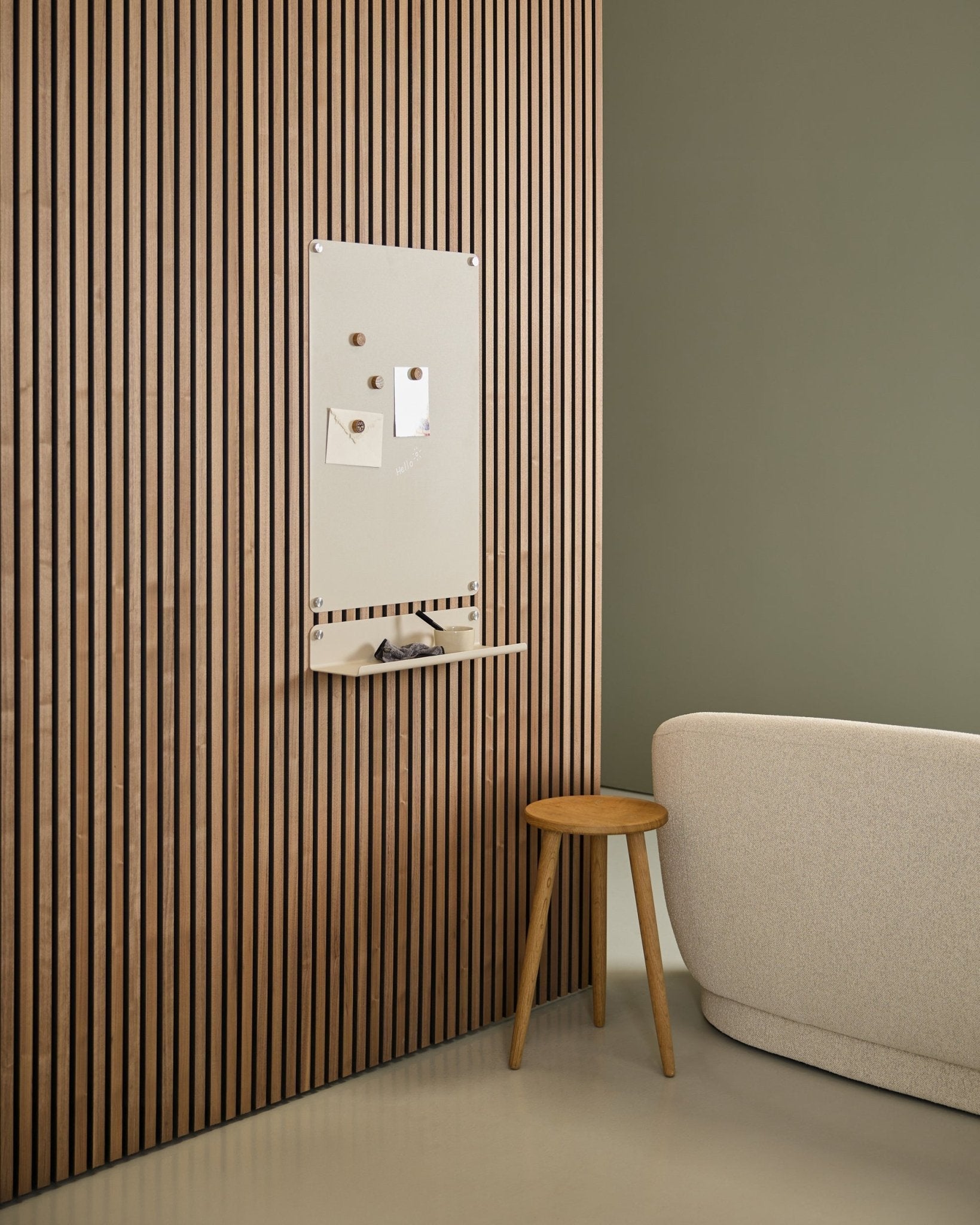 Minimalistisk interiør med beige sofa og krakk i tre, med en large beige WoodUpp Norr magnettavle montert på et nordisk Akupanel akustisk trepanel nær en grønn vegg.