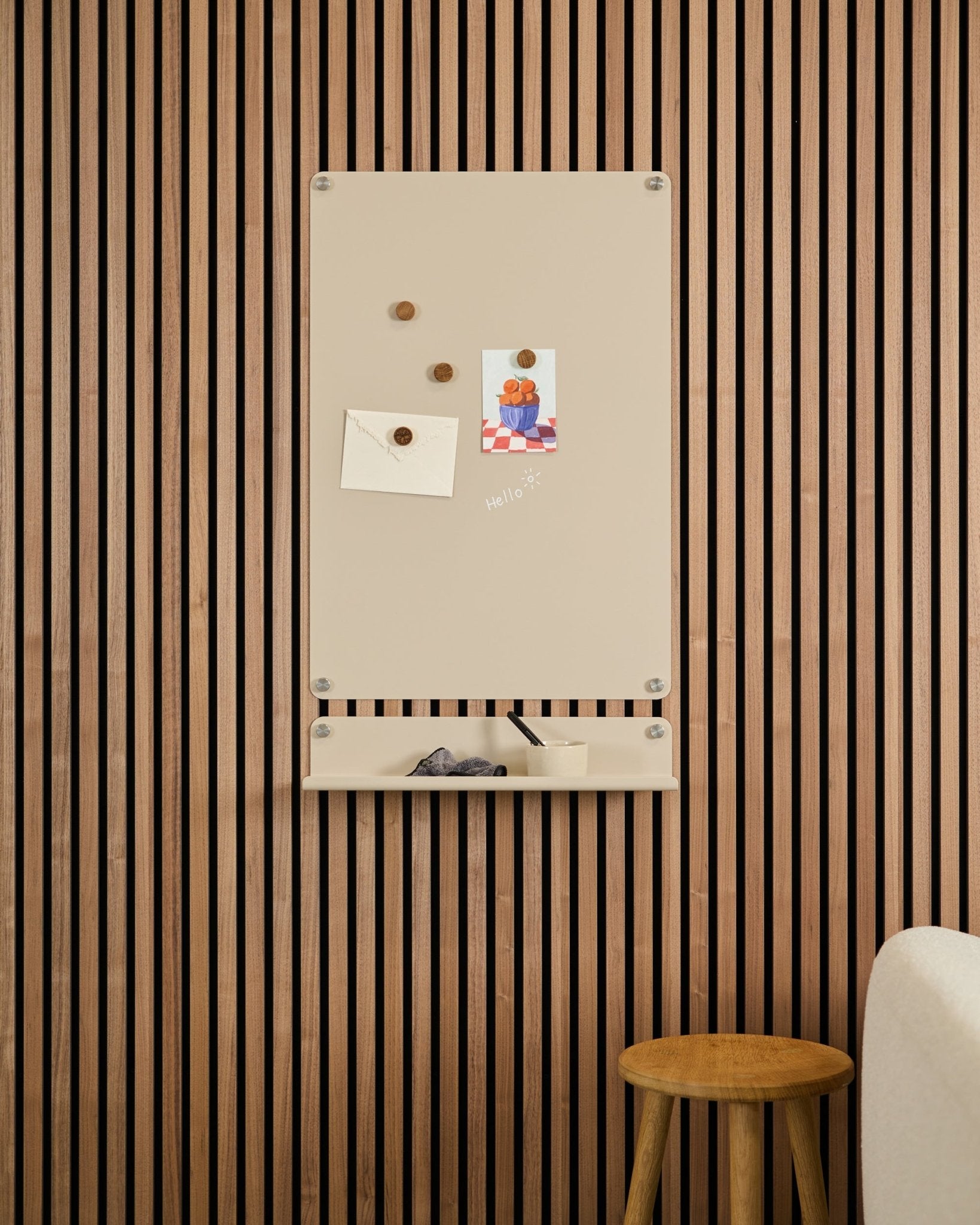 Beige Norr magnettavle fra WoodUpp montert over en small hylle på en vegg med Akupanel akustiske trepaneler, med en krakk og en hvit sofa i nærheten.