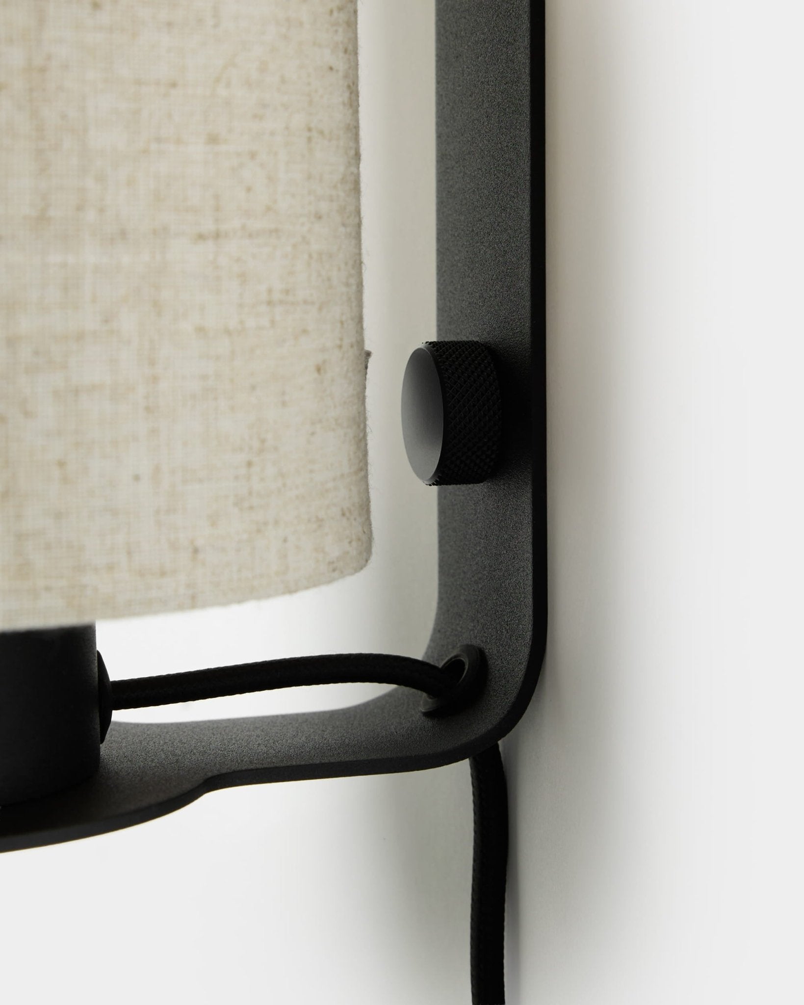 Nærbilde av WoodUpp Feya vegglampe i black og beige, med teksturert bryterknott, skjerm i lin-bomull og synlig kabel montert på en hvit vegg.