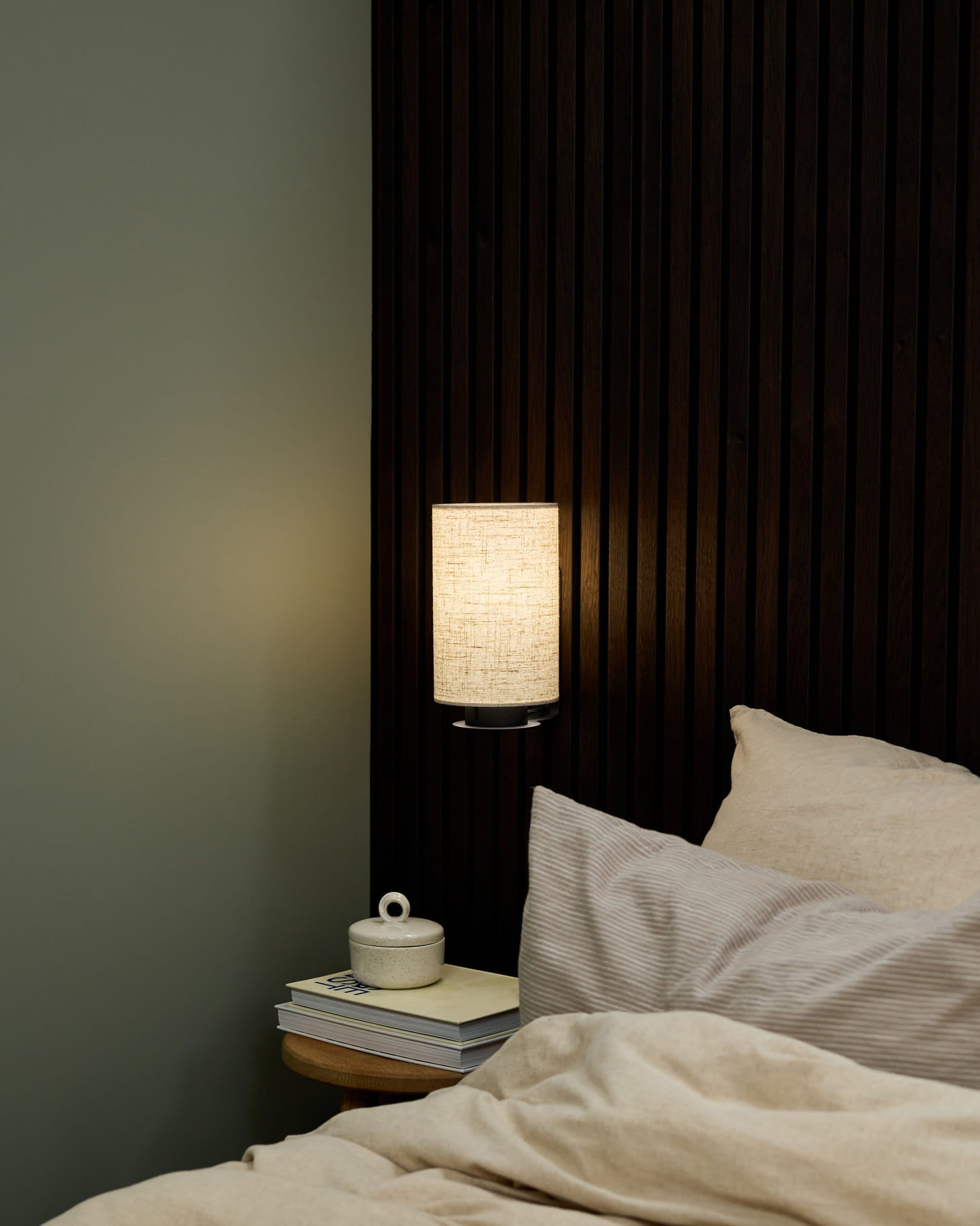 Soveromshjørne med en WoodUpp Feya vegglampe i Black/Beige, sengetøy i beige og et nattbord med bøker og en keramisk kopp med lokk.