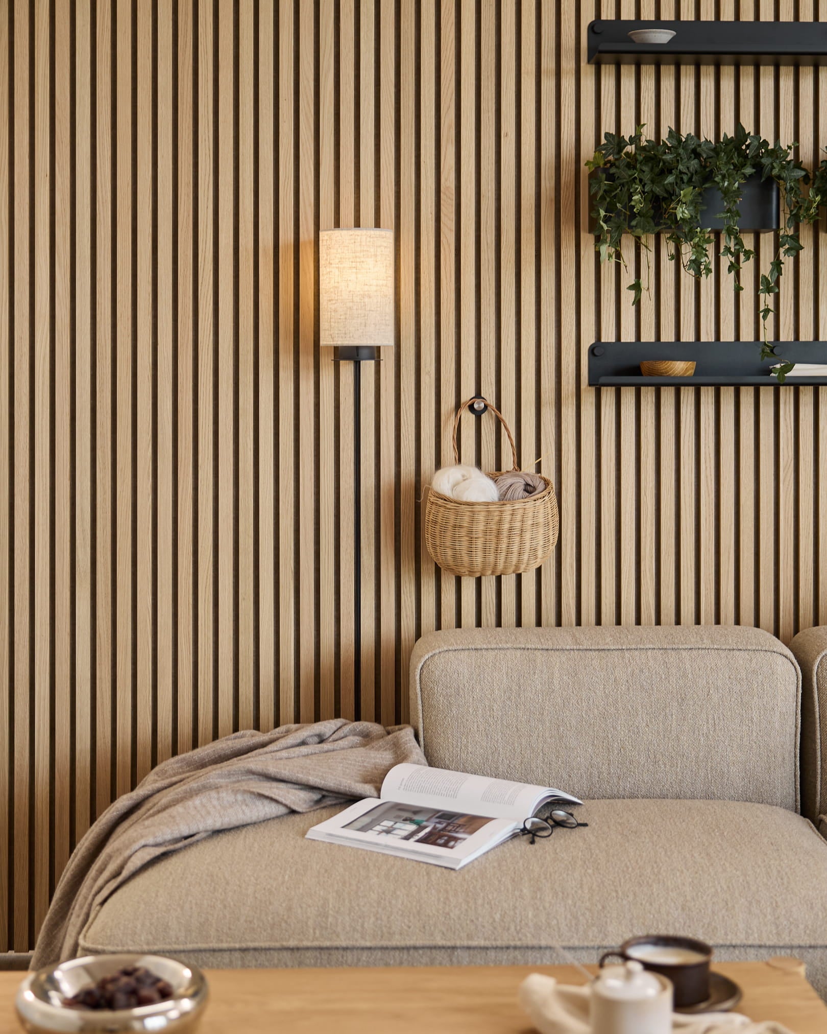 Sofa i Beige med pledd og bok ved siden av en WoodUpp akustisk spilevegg i Akupanel, hyller med planter og dekor, og en Feya vegglampe i Black/Beige.