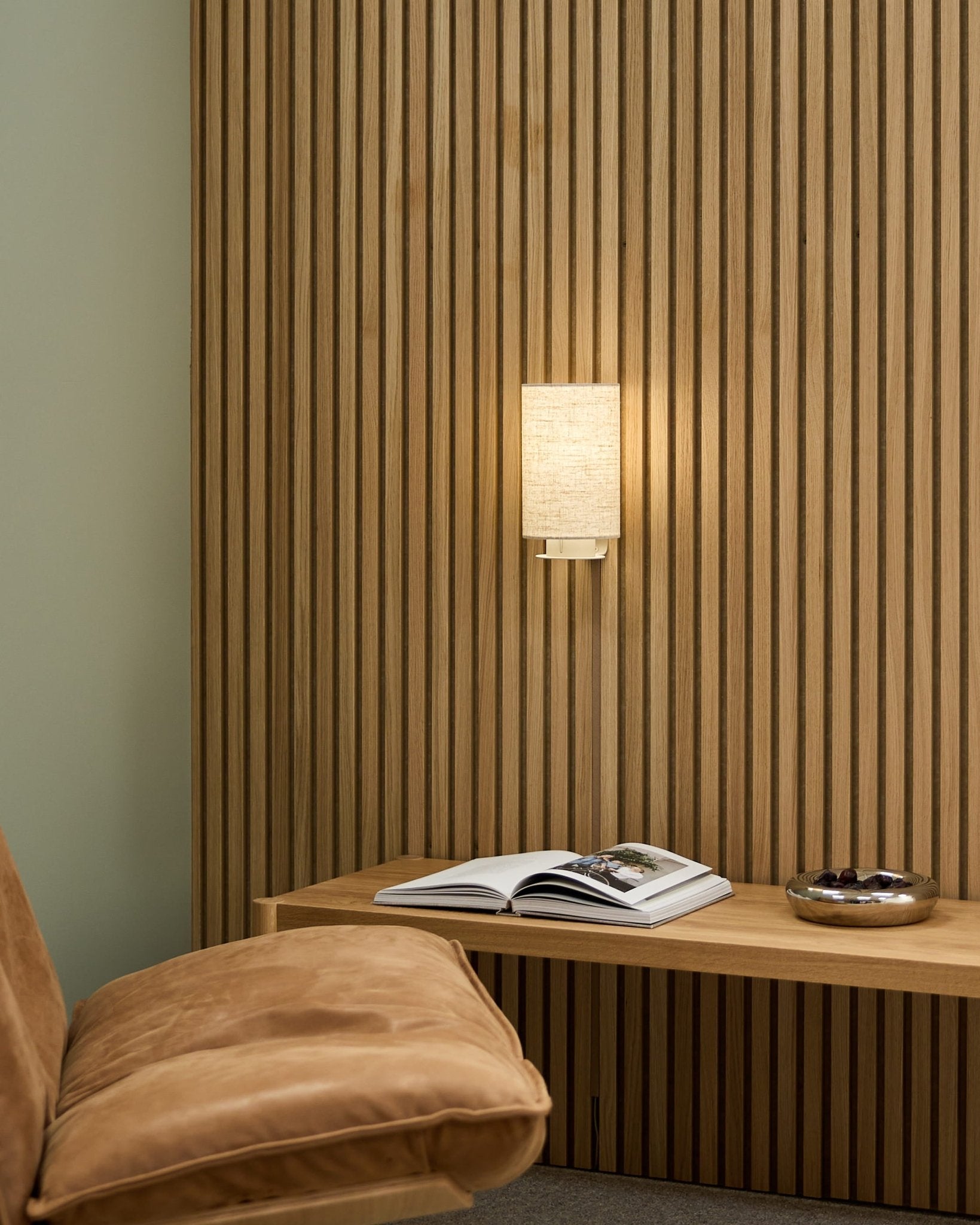 Minimalistisk nordisk rom med en lys brun stol, WoodUpp Akupanel akustisk spilevegg, Feya vegglampe i Beige/Beige, en åpen bok på en benk og en skål.
