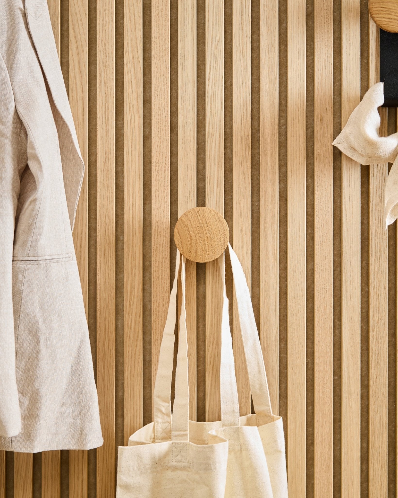 En beige kåpe og handlenett henger på Linn I Single Hanger i Natural Oak fra WoodUpp, montert på et vertical Akupanel akustikkpanel i trespiler.