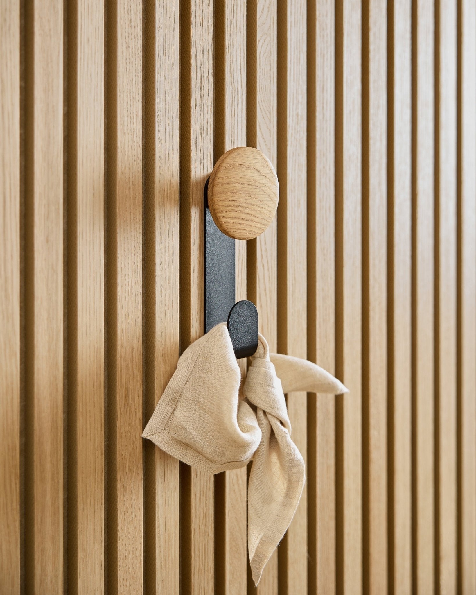 Et beige klede henger fra Linn Vertical Hanger i Natural Oak fra WoodUpp, montert på et vertical Akupanel akustisk spilepanel.
