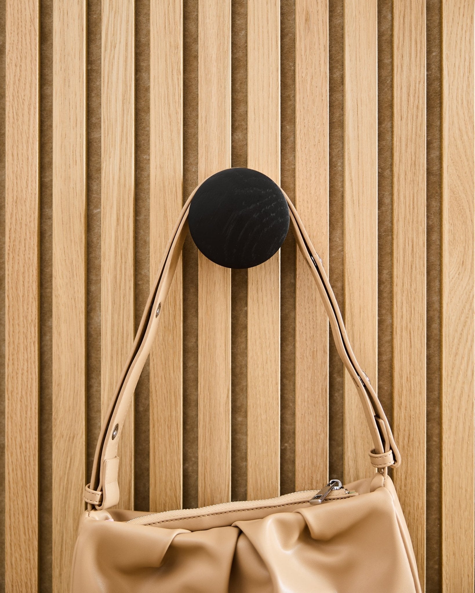 Beige veske henger på en WoodUpp Linn | Single Hanger i Black Oak, montert på et akustisk spilepanel i lyst tre Akupanel.