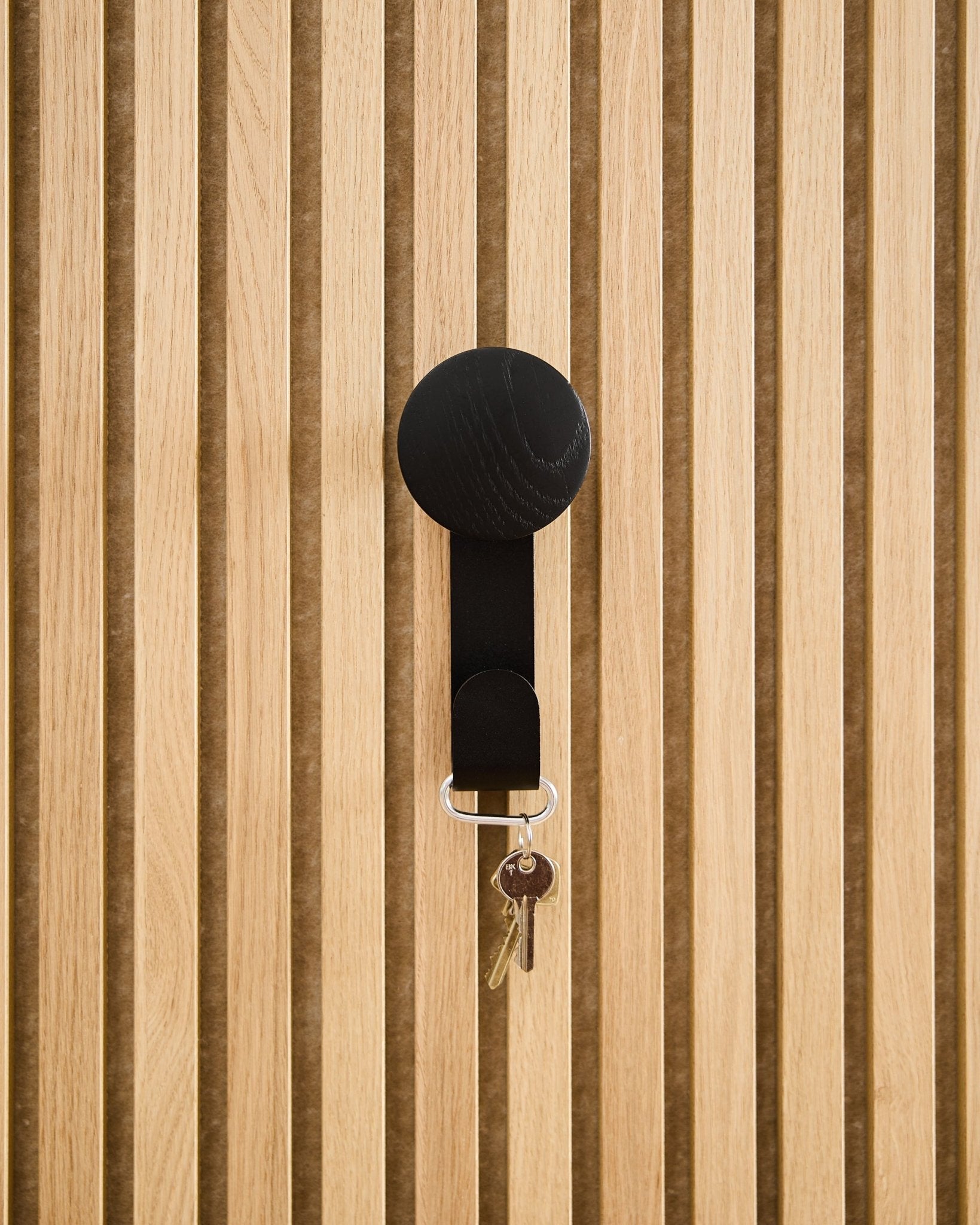 En Linn Vertical Hanger i Black Oak fra WoodUpp holder en nøkkelring med to nøkler og en rund brikke, montert på et teksturert Akupanel akustisk trepanel.