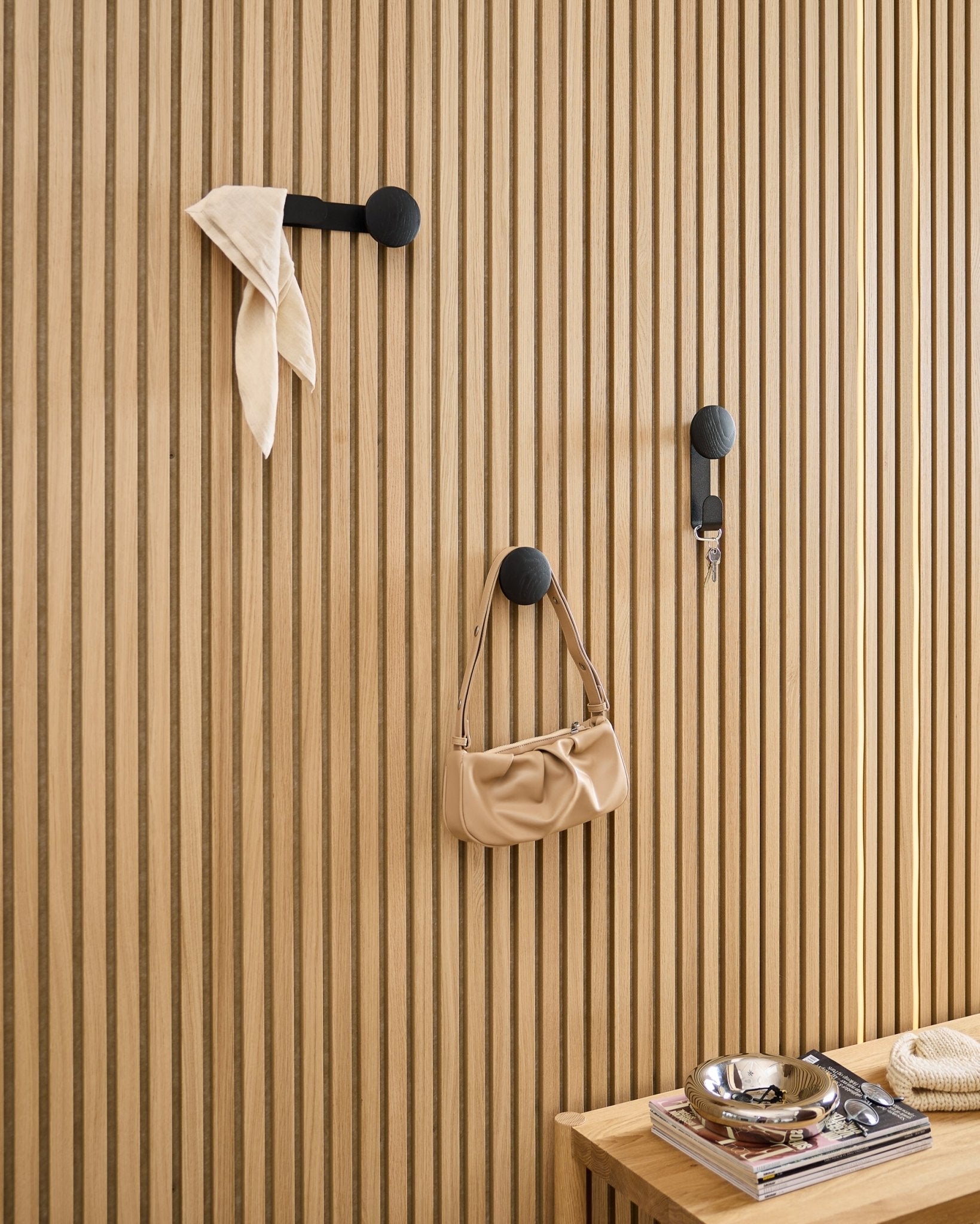 WoodUpp Linn Single Hanger i Black Oak holder et skjerf, en beige veske og nøkler på et black Akupanel akustisk trepanel; benk med bøker og metallfat i nærheten.