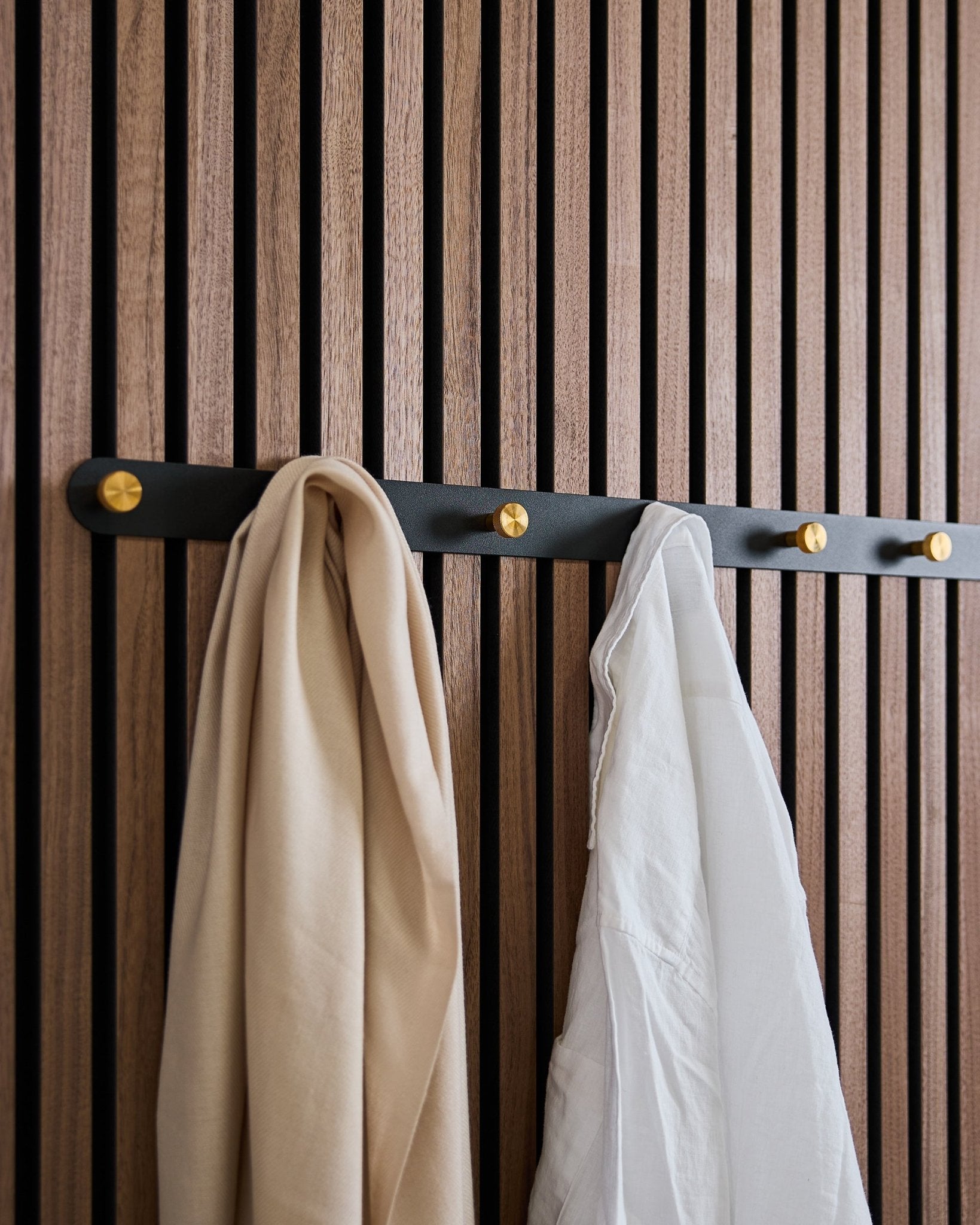 WoodUpp Norr Coat Hook Rack i black/brass (X7) med et beige skjerf og en hvit skjorte henger på et nordisk akustisk spilepanel i tre.