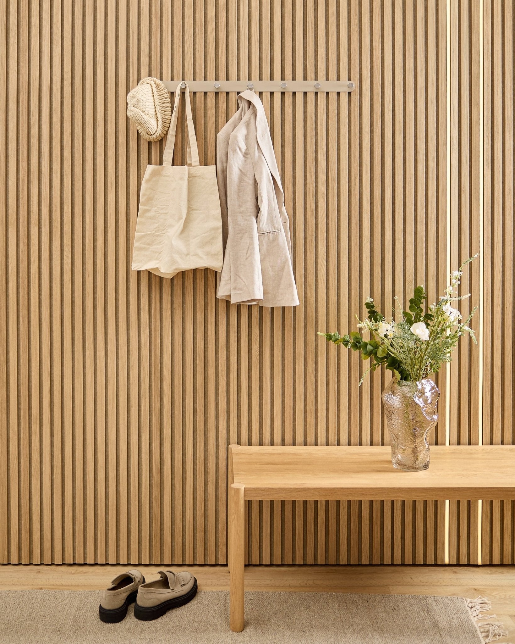En beige veske, lue og skjorte henger på WoodUpp Norr knaggrekke over sko, med en benk og vase ved siden av det stripete akustiske spilepanelet.