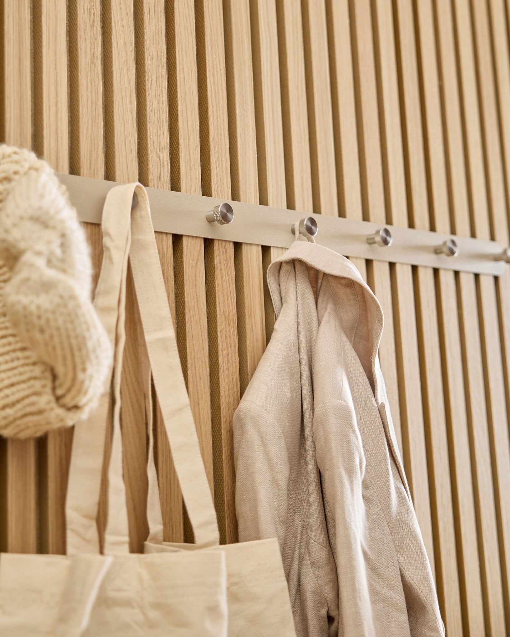 En beige totebag, strikkelue og linskjorte henger på WoodUpp Norr knaggrekke (Beige/rustfritt Steel, X7) montert på et lyst spilepanel i tre Akupanel.