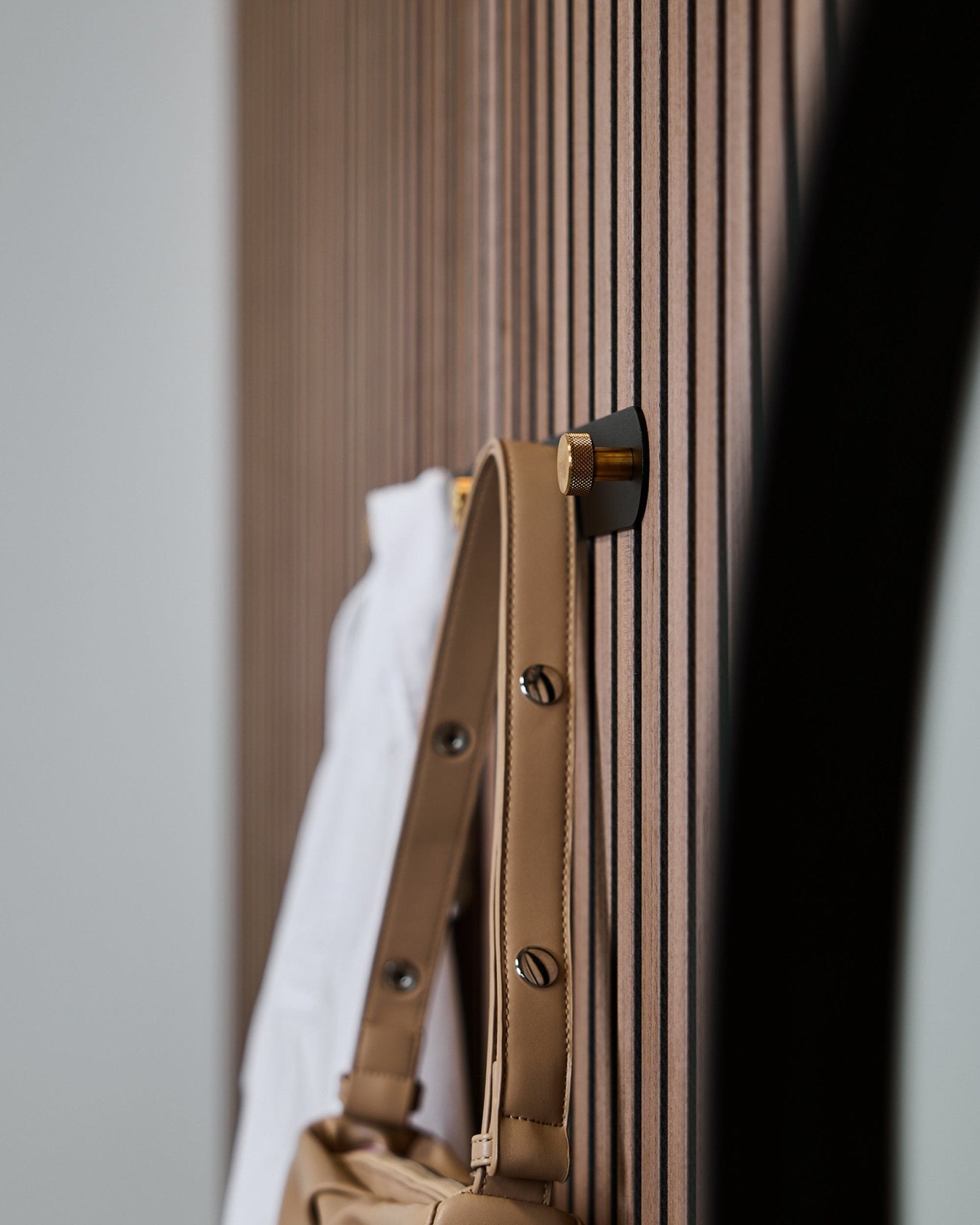 En lys brun veske og en hvit skjorte henger på en WoodUpp Norr knaggrekke (Black/Brass, X5) montert på et vertical Akupanel akustisk spilepanel.