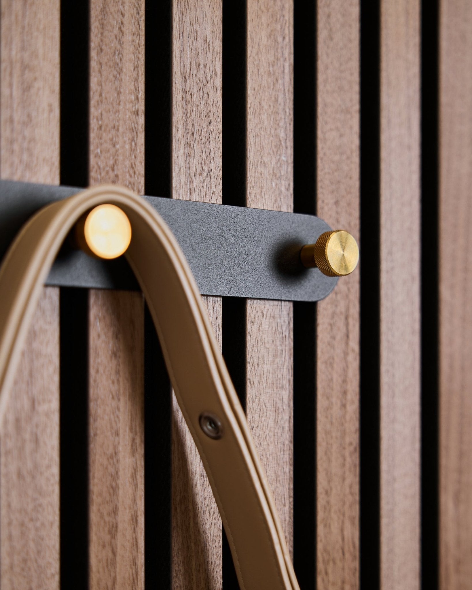 Fem black/brass knagger på Norr knaggrekke fra WoodUpp er montert på et akustisk spilepanel i tre, og hver av dem holder en beige stropp.