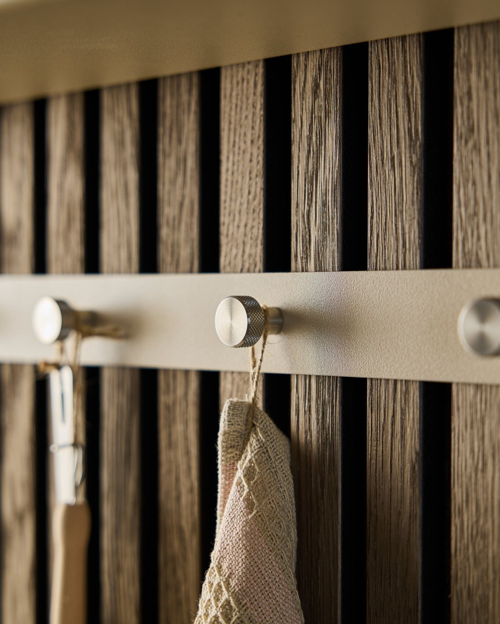 Nærbilde av WoodUpp Norr knaggrekke (Beige/rustfritt Steel, X5) montert på en vegg med trespiler Akustikkpanel, med et håndkle og et verktøy med håndtak hengende på.