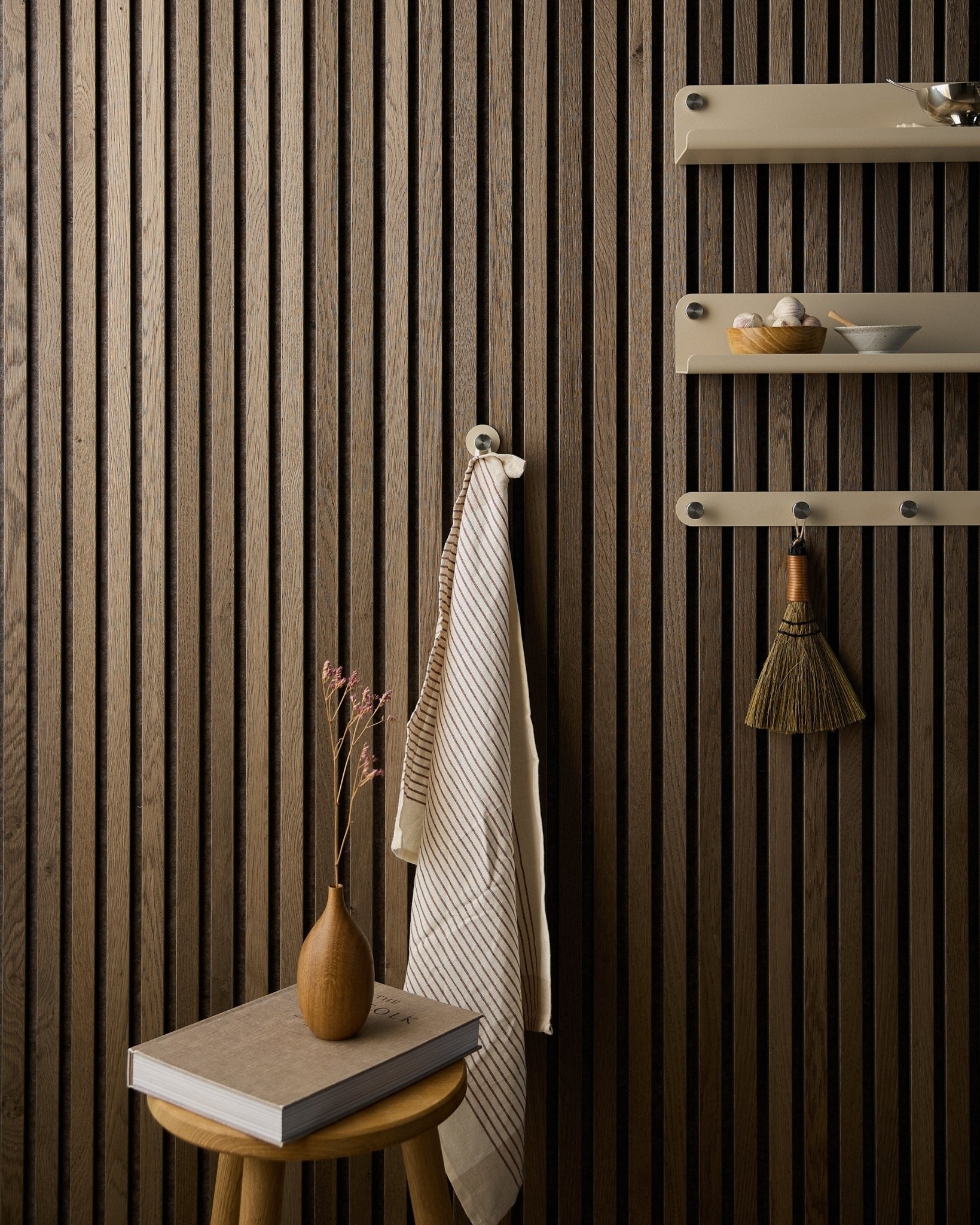 Et håndkle henger på en Norr knagg fra WoodUpp, montert på en Akupanel akustisk spilevegg i tre ved siden av beige hyller, med en kost, krakk, vase og bok.