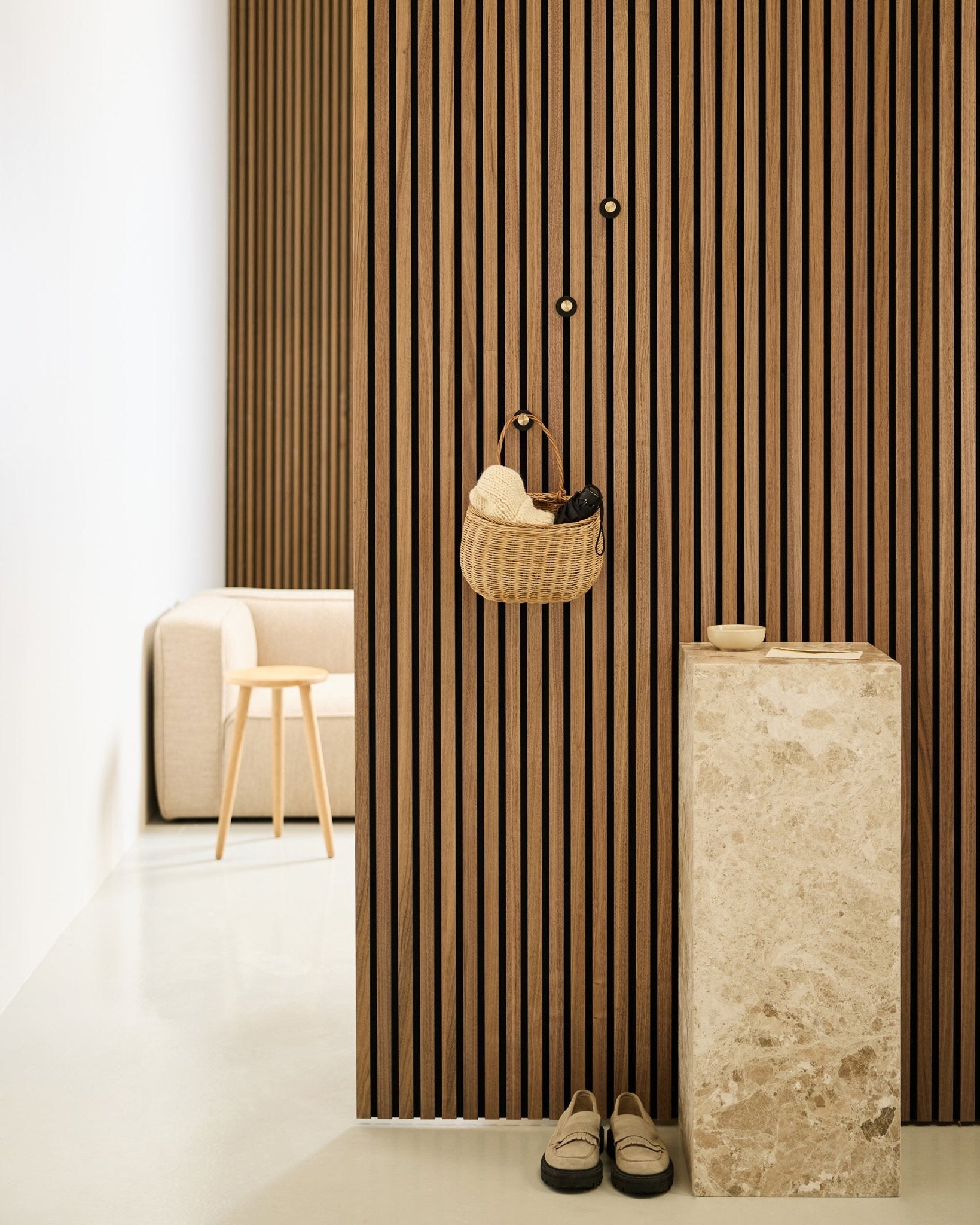 Minimalistisk entré med Akupanel akustisk spilevegg, WoodUpp Norr Coat Hook i black og brass, kurv, sokkel i stein, tøfler og beige stol.