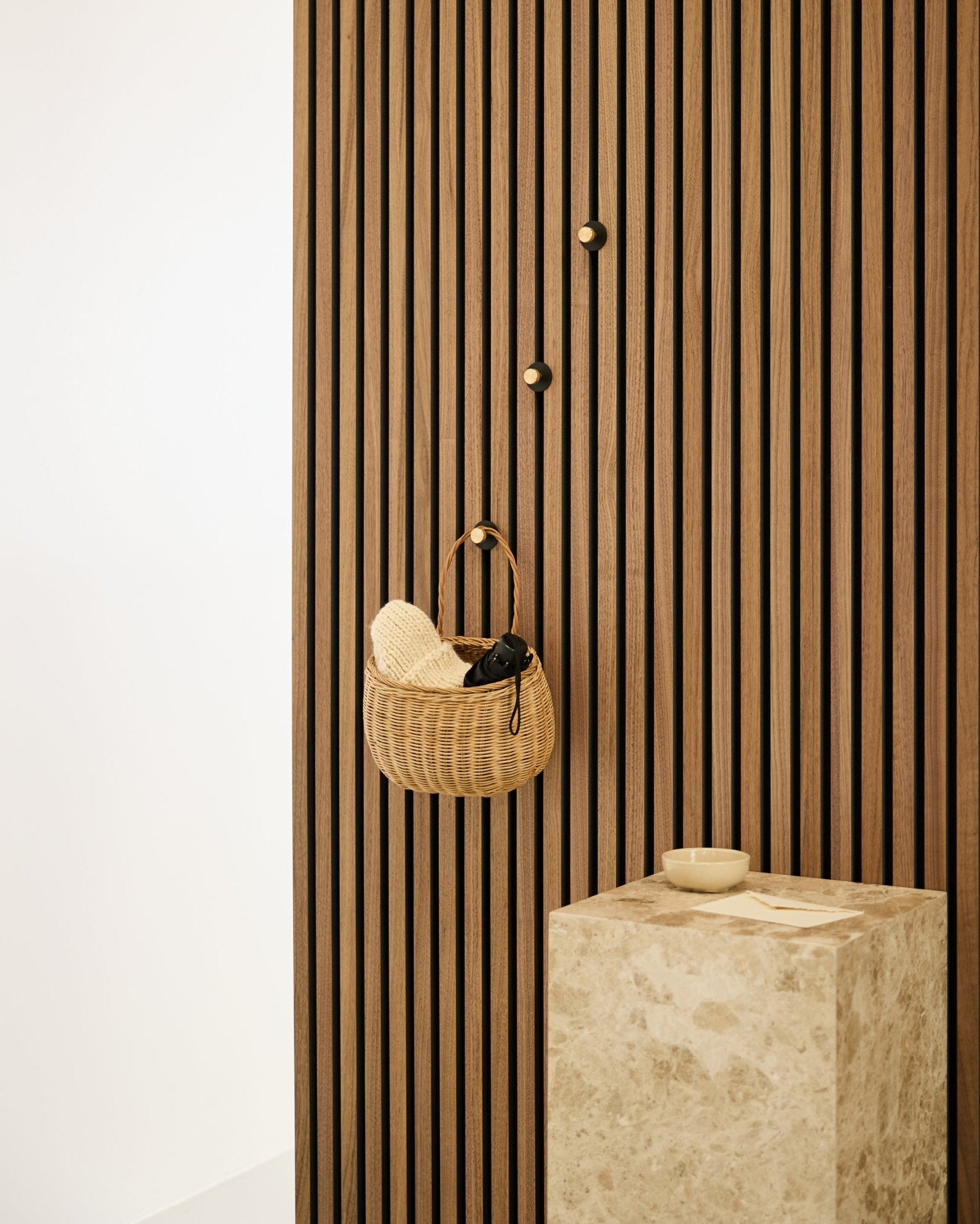 Norr Coat Hook fra WoodUpp montert på en nordisk Akupanel akustisk spilevegg, ved siden av en beige sokkel i stein med en small skål i et minimalistisk interiør.